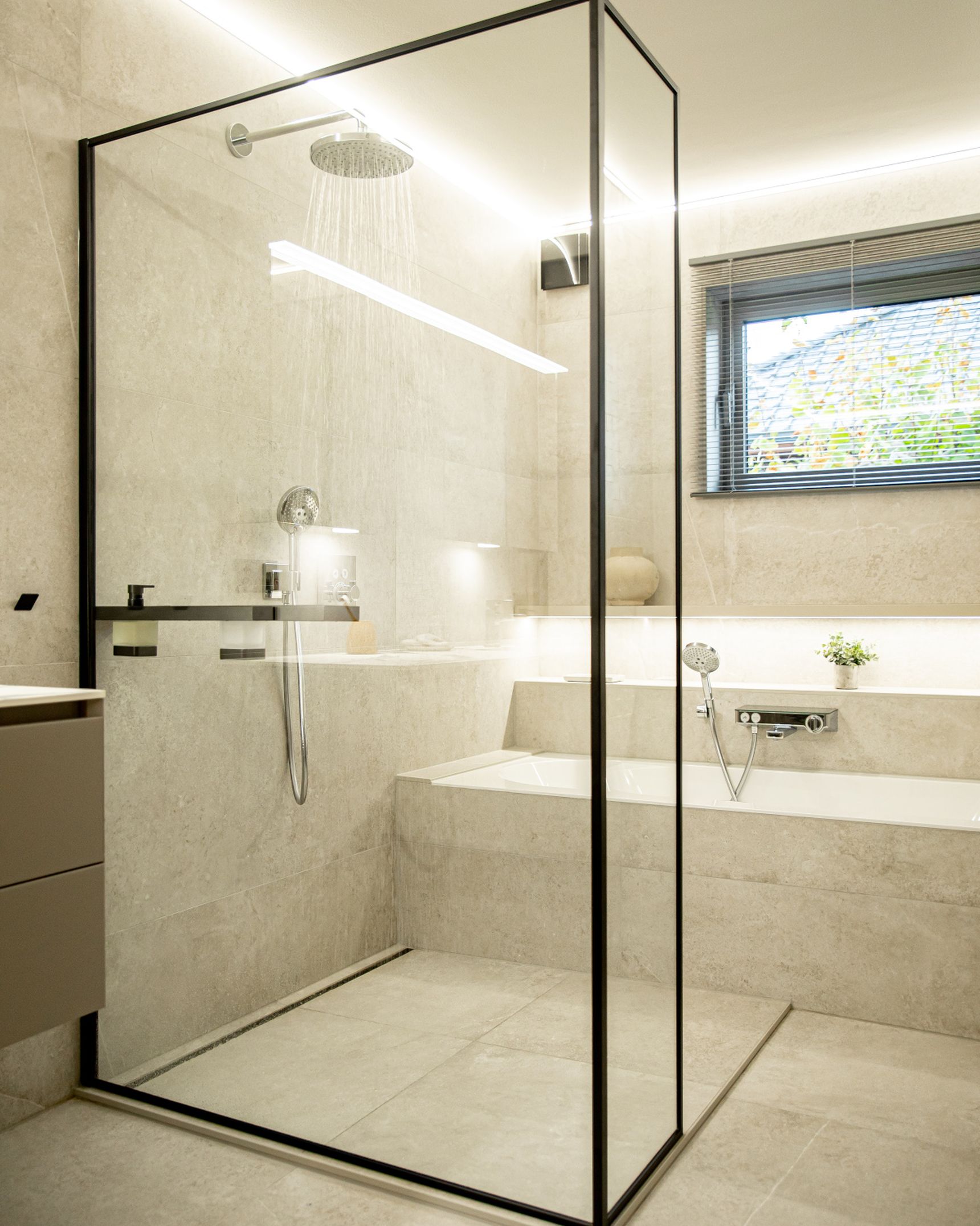Modern bathroom with a glass shower enclosure, rainfall showerhead, beige tiles, bathtub, and a window with blinds.