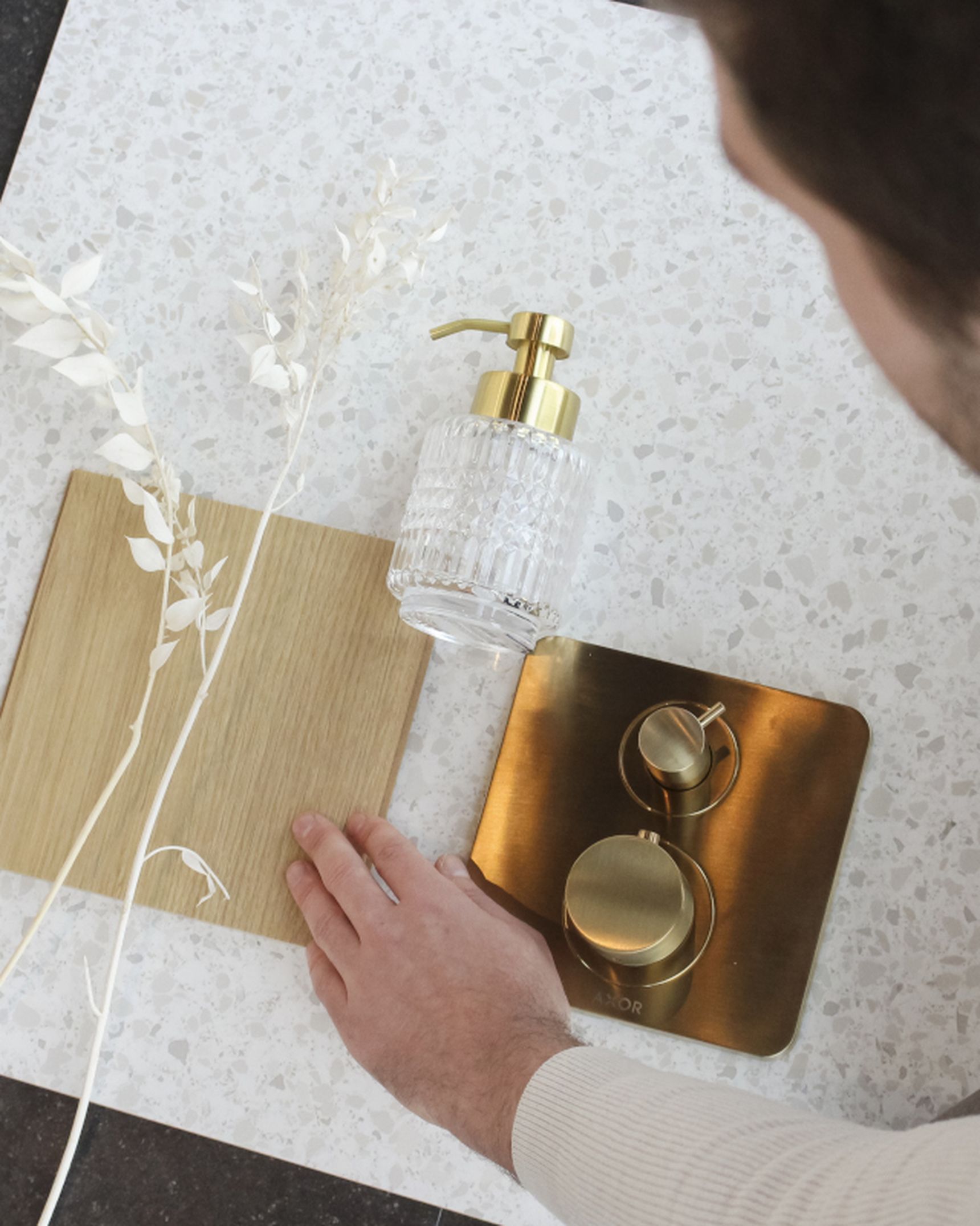 "Man rangschikt decoratieve items zoals een houten tegel, droogbloemen, glazen zeeppompje en messing kraan op een terrazzo-oppervlak.