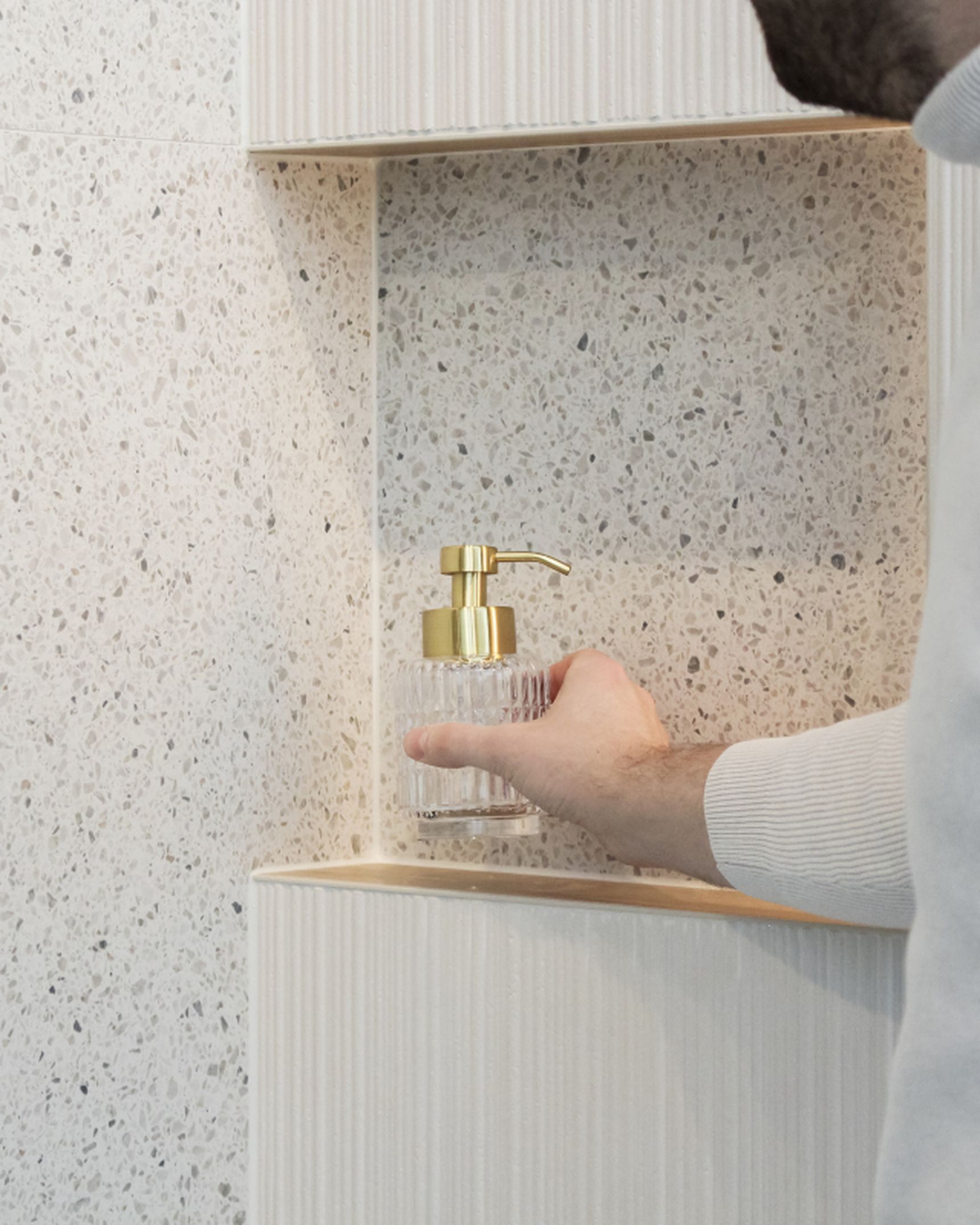 Persoon in wit hemd reikt naar een glazen zeeppompje met gouden pomp op een gespikkeld badkamerrek.