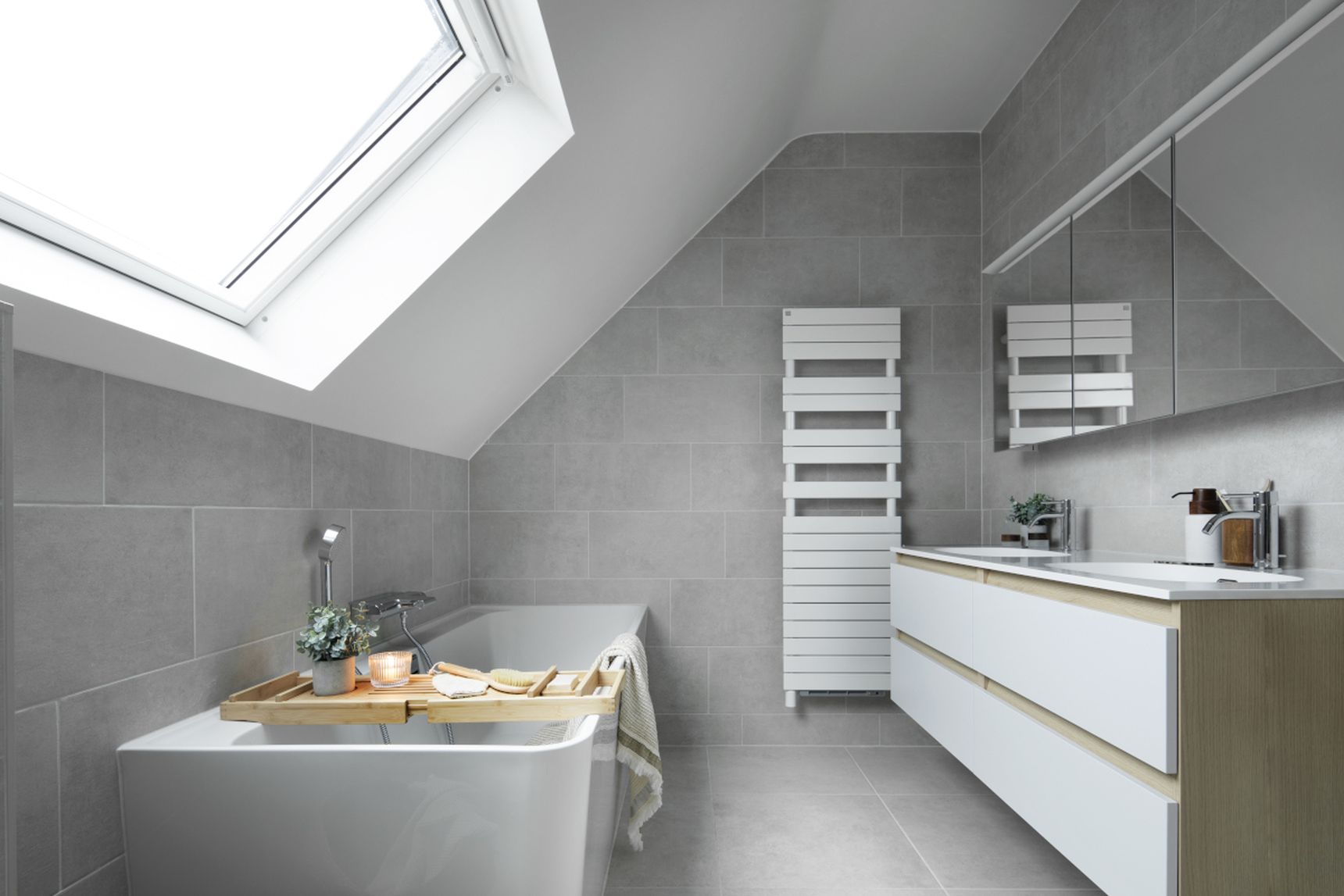 Modern bathroom with a skylight, gray tiles, a bathtub with a wooden tray, a towel, and a vanity with a mirror and sink.