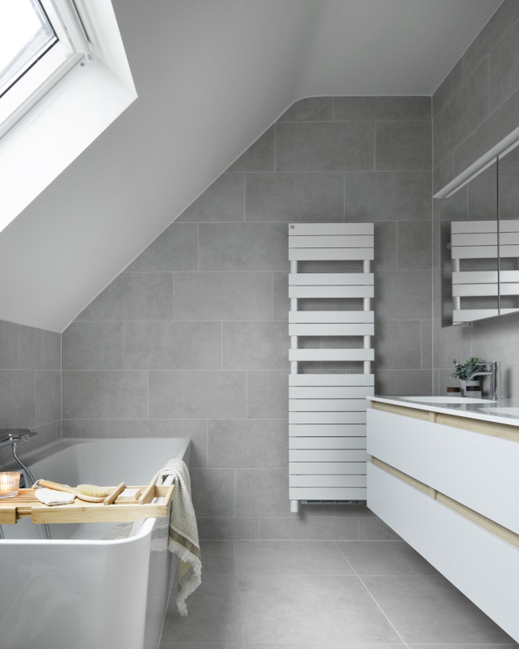 Moderne badkamer met dakraam, grijze tegels, bad met houten plank en handdoek, en een badkamermeubel met spiegel en wastafel.
