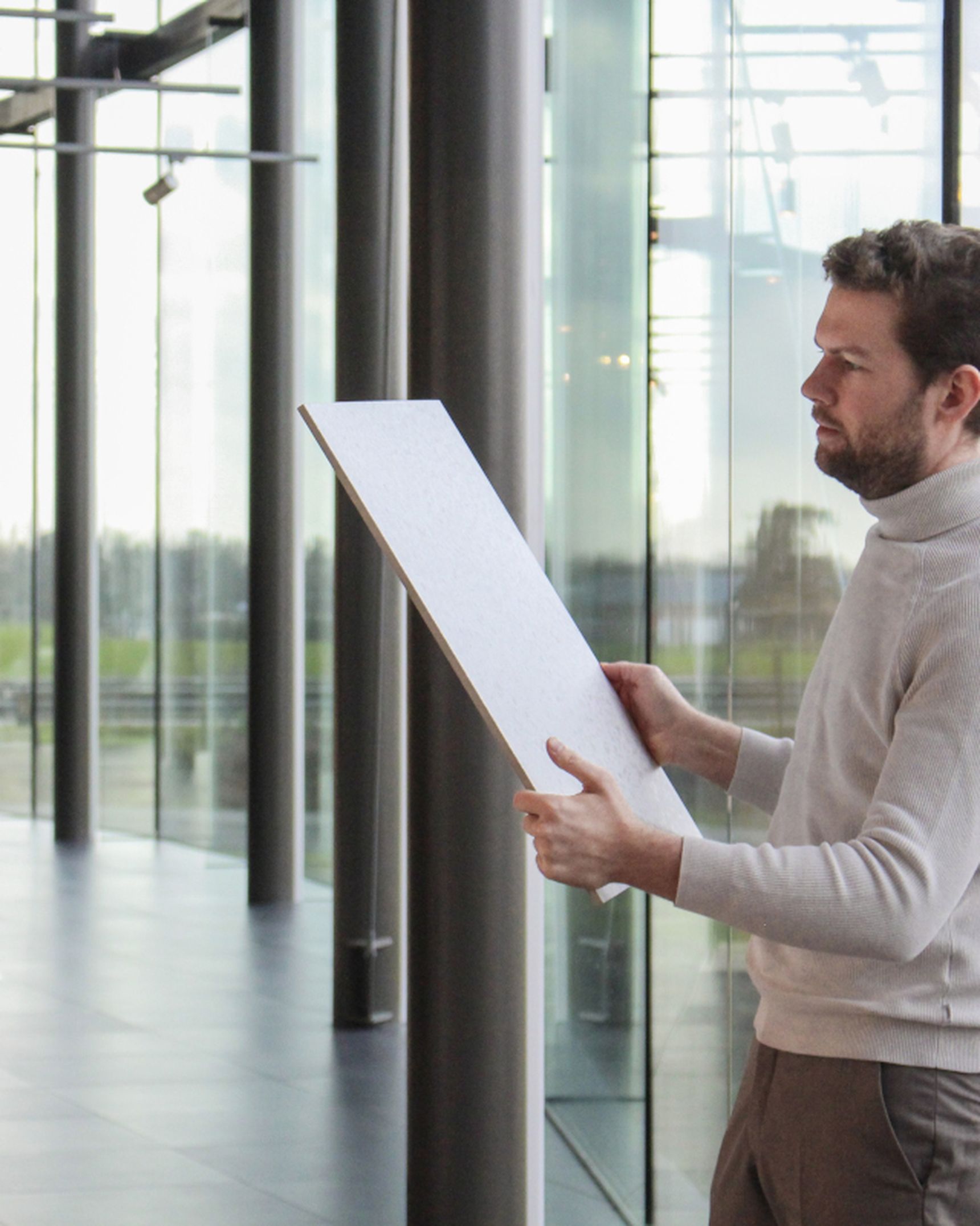 Pieter Steylaerts bekijkt een grote tegel in een moderne toonzaal van Steylaerts met glazen wanden en zicht op de buitenomgeving.