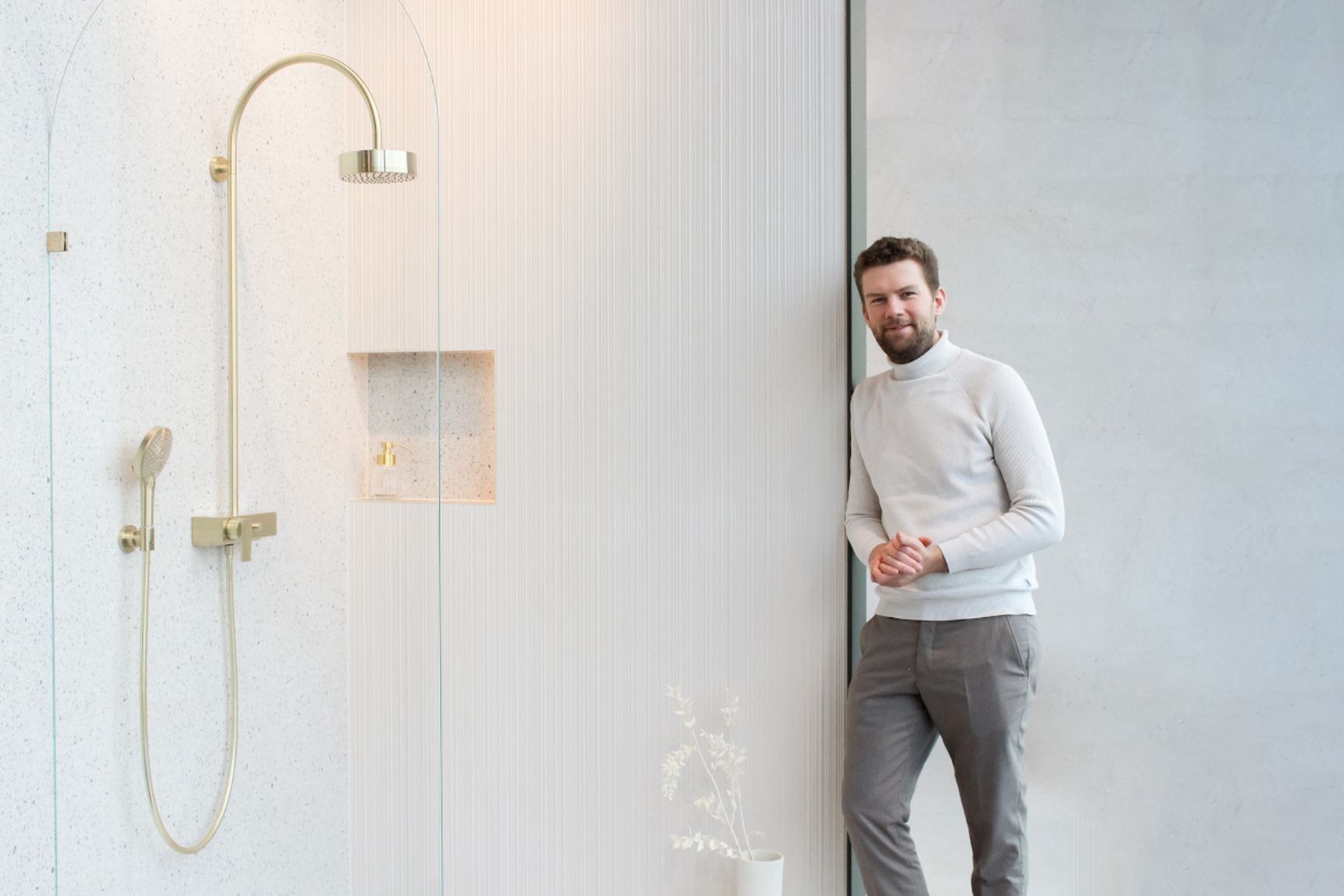 Pieter Steylaerts in witte trui en grijze broek naast moderne douche met gouden kranen in een minimalistische badkamer.