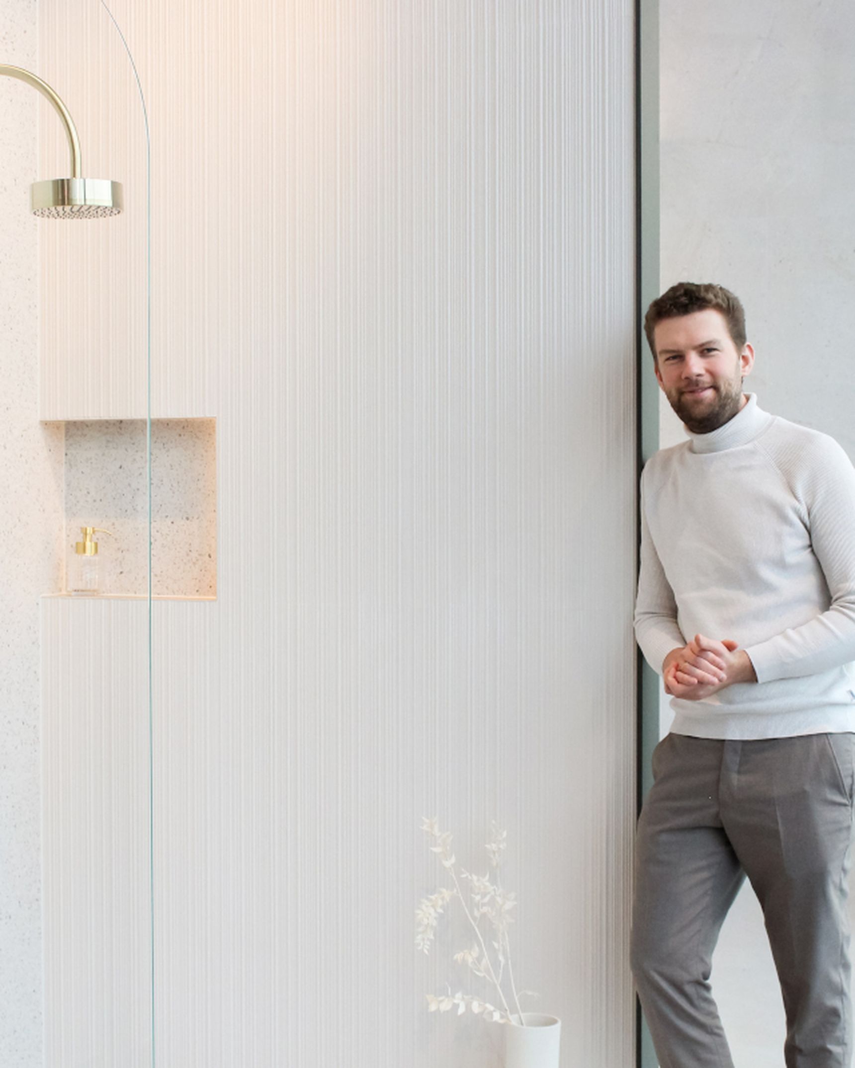 A man in a white sweater and gray pants stands beside a modern shower with gold fixtures in a minimalist bathroom.