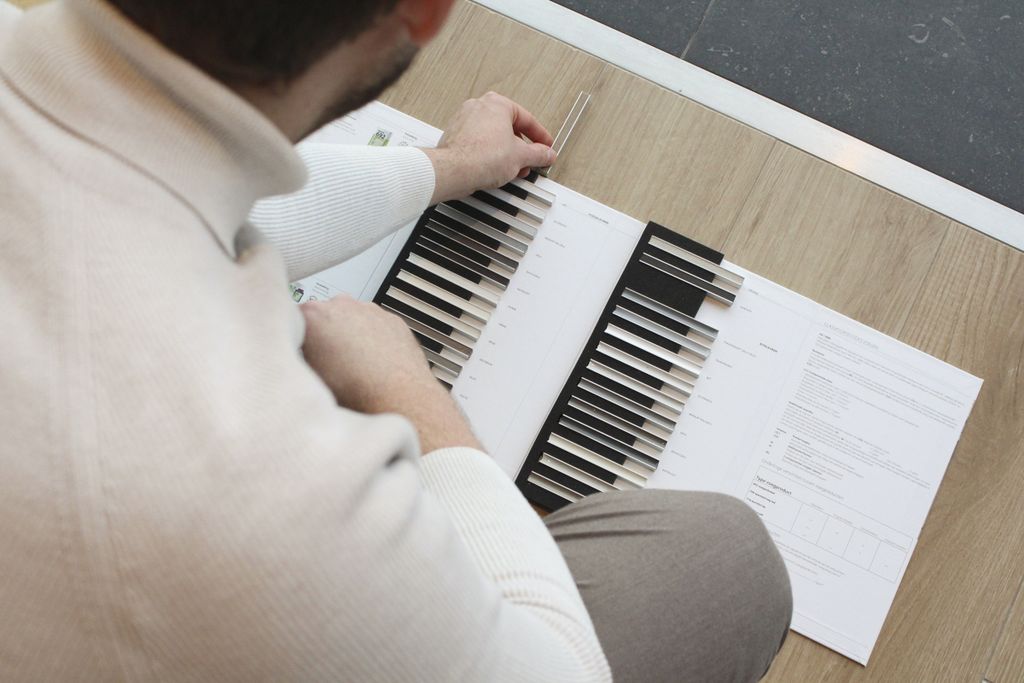 Person examining a color swatch book on a wooden floor, with documents nearby. They are wearing a white sweater and gray pants.