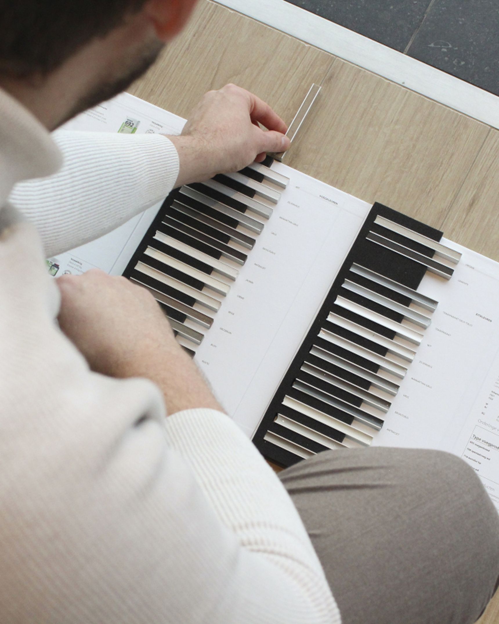 Persoon in witte trui en grijze broek bekijkt een kleurenstaalboek op een houten vloer, met documenten in de buurt.