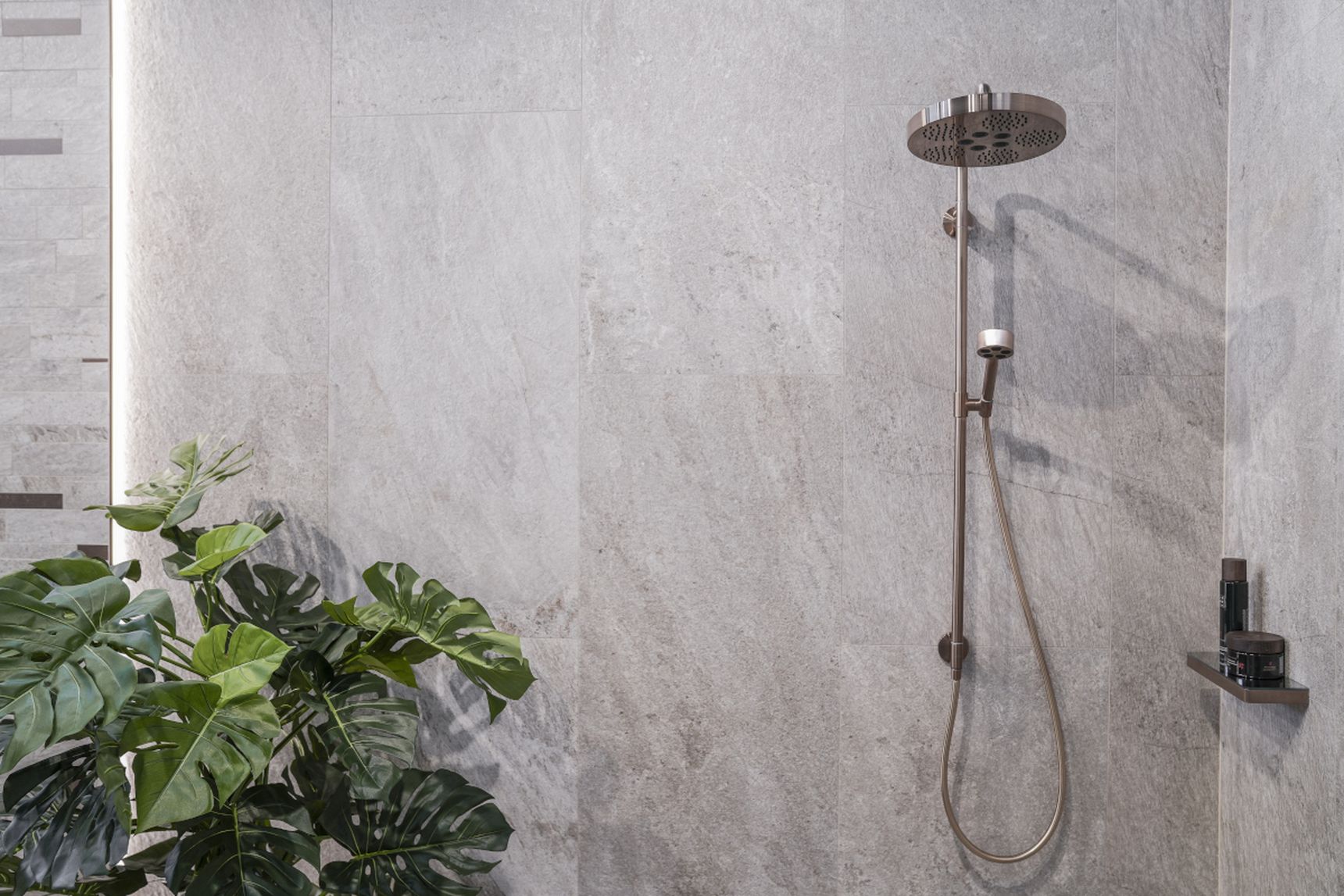 Modern shower with a rainfall showerhead and handheld sprayer on gray tiled wall, next to a green plant and small shelf with toiletries.