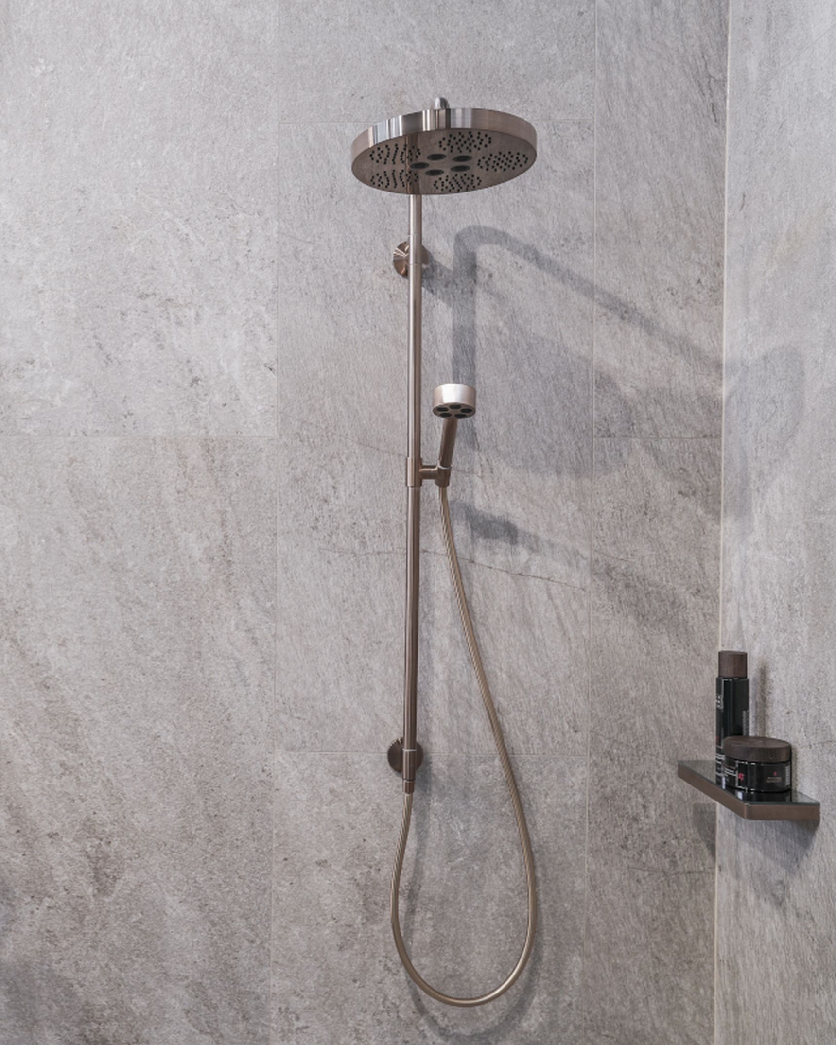 Modern shower with a rainfall showerhead and handheld sprayer on gray tiled wall, next to a green plant and small shelf with toiletries.