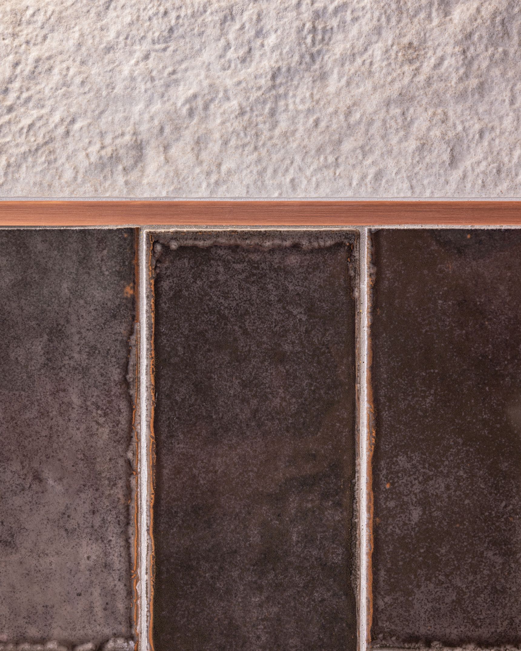 Detail van keramische wandtegels (Sunrise Grey Strutt en Sunrise Grey Plissé Mix) met glanzende, donkere afwerking en een smal profiel als overgang naar een lichte, gestructureerde tegel erboven.