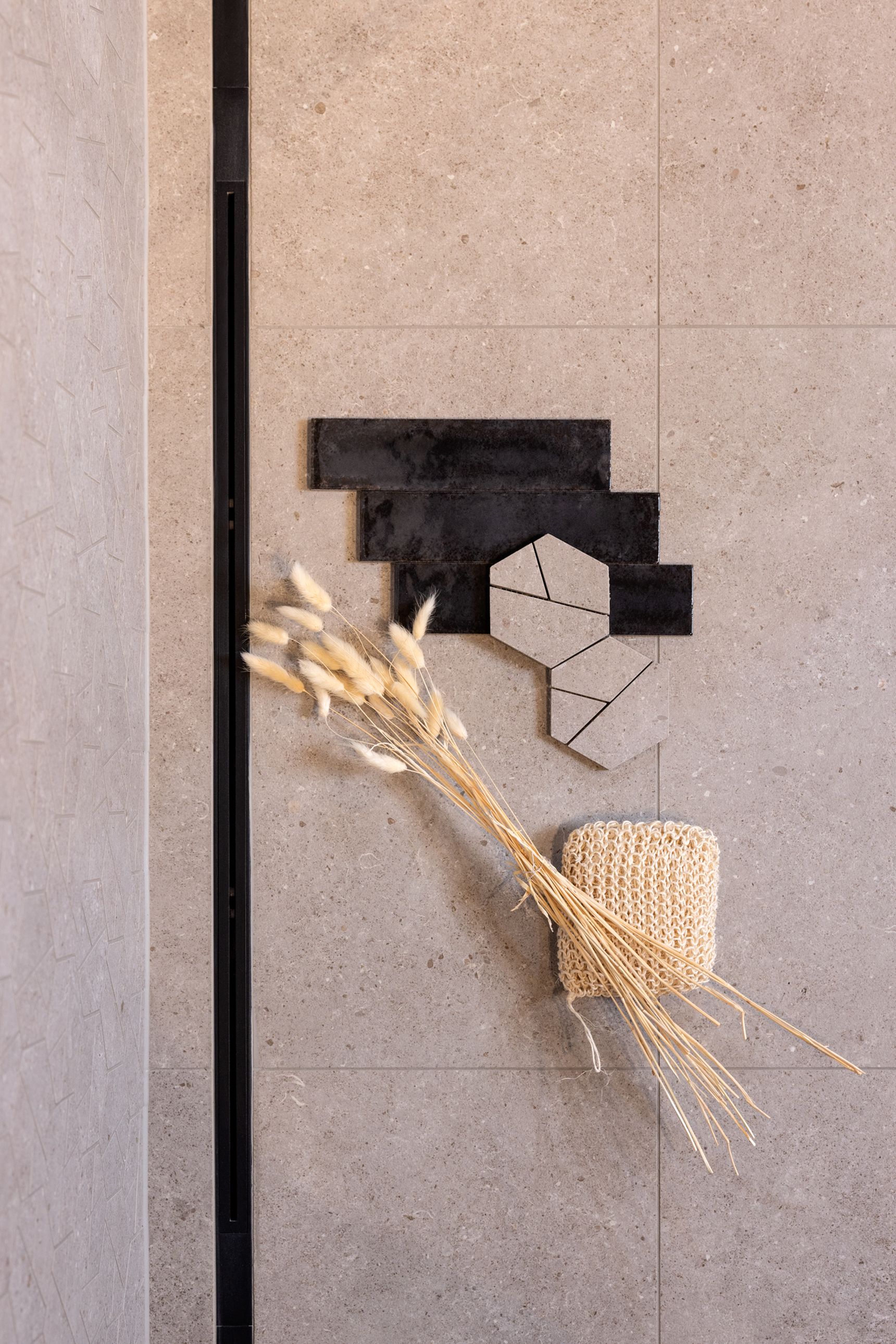 Minimalistische douchewand met beige natuursteenlook tegels, een zwarte inbouwnis, een geometrisch tegelaccent in zwart en beige, een bundel gedroogde siergrassen en een natuurlijke spons.
