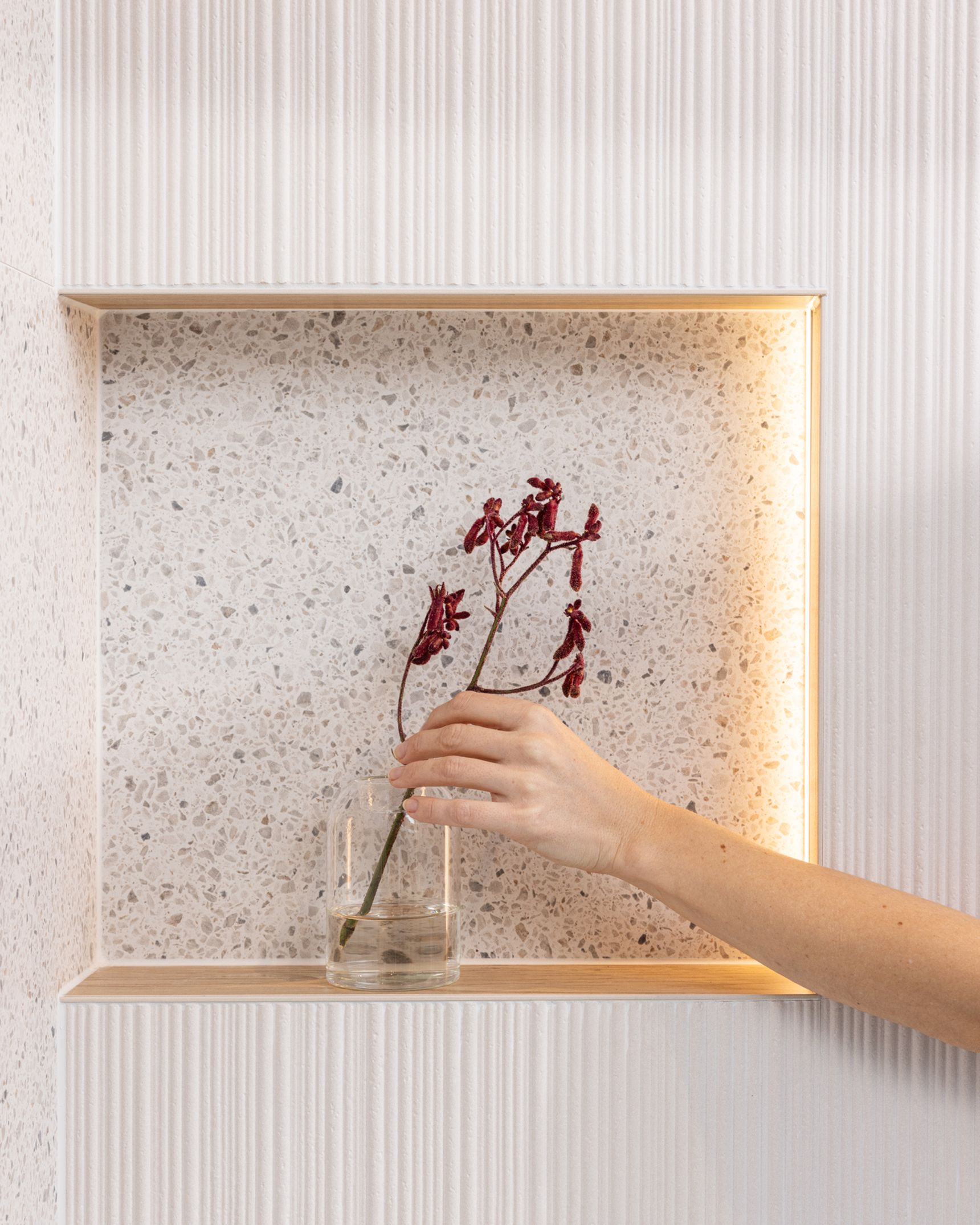 Hand plaatst glazen vaas met rode bloemen op een verlichte nisplank in een wand met gespikkeld oppervlak.