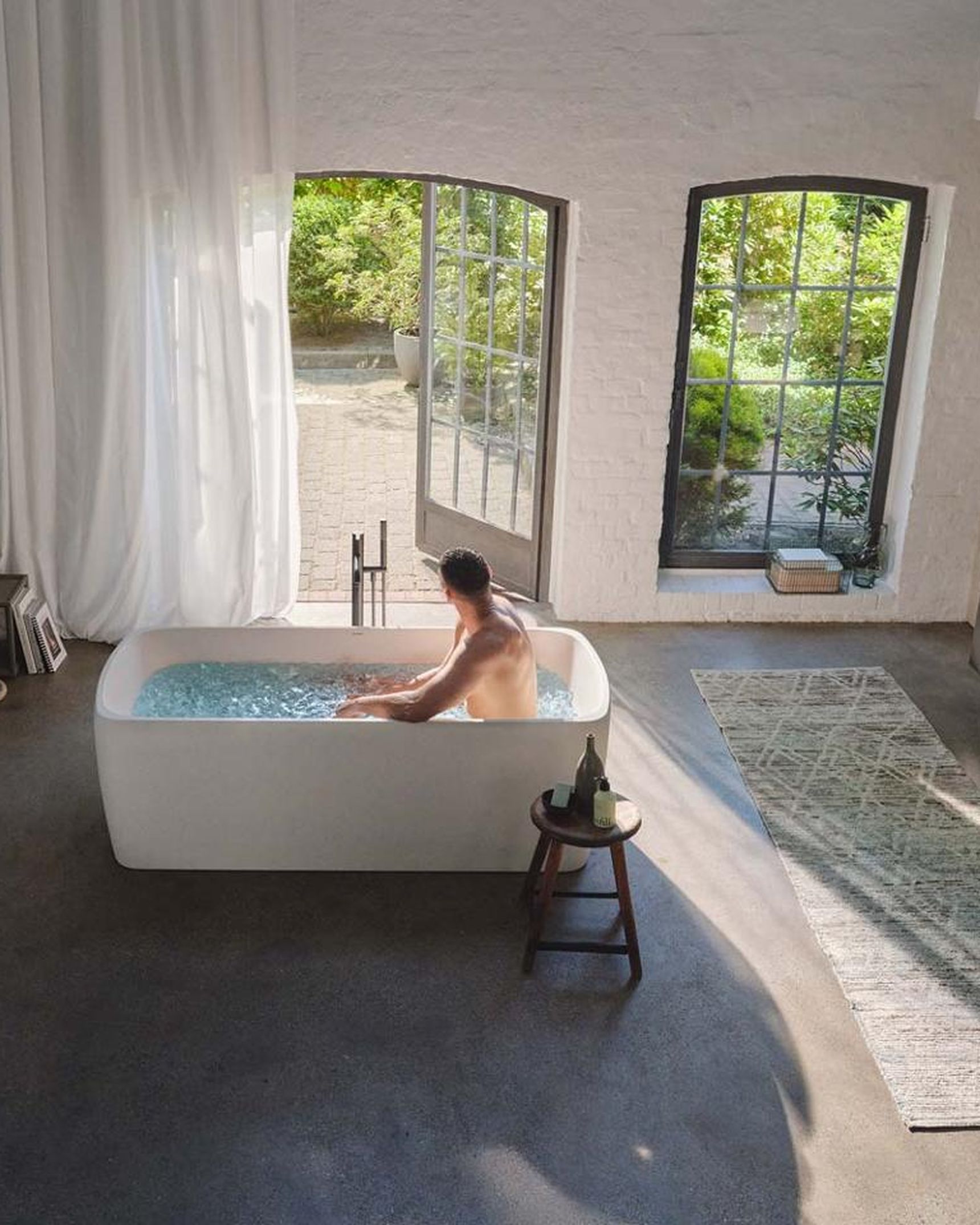 Ruime badkamer met vrijstaand champagnebubbelbad, waarin een man ontspant, omgeven door grote ramen en een moderne inrichting.