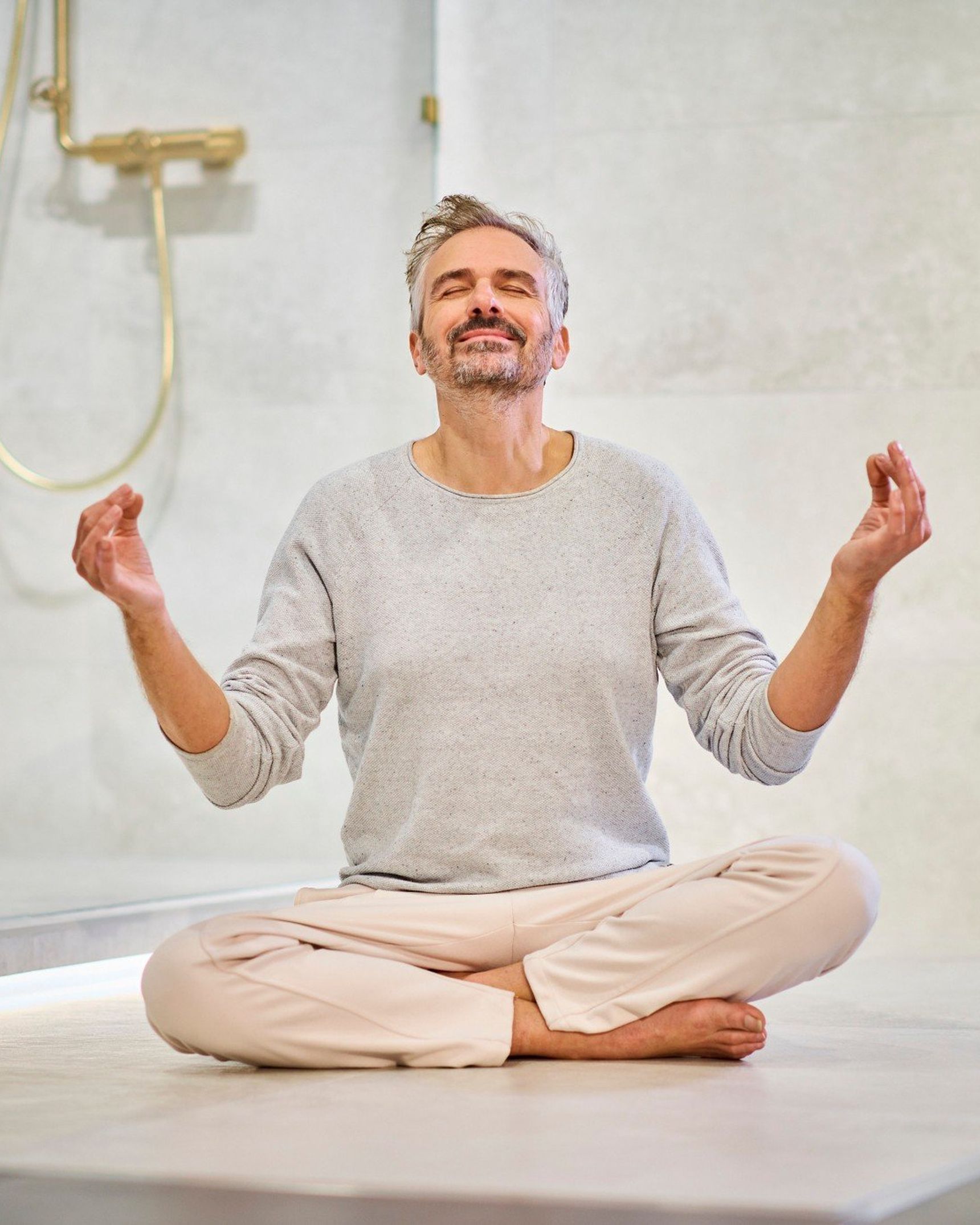 A man sits cross-legged, meditating with a serene expression in a modern room with a shower and greenery on the wall.