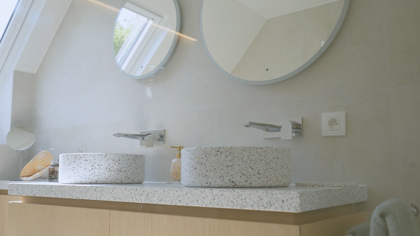 Modern bathroom with two round mirrors, terrazzo sinks, and sleek faucets on a wooden vanity against a light tiled wall. Natural light from skylight.
