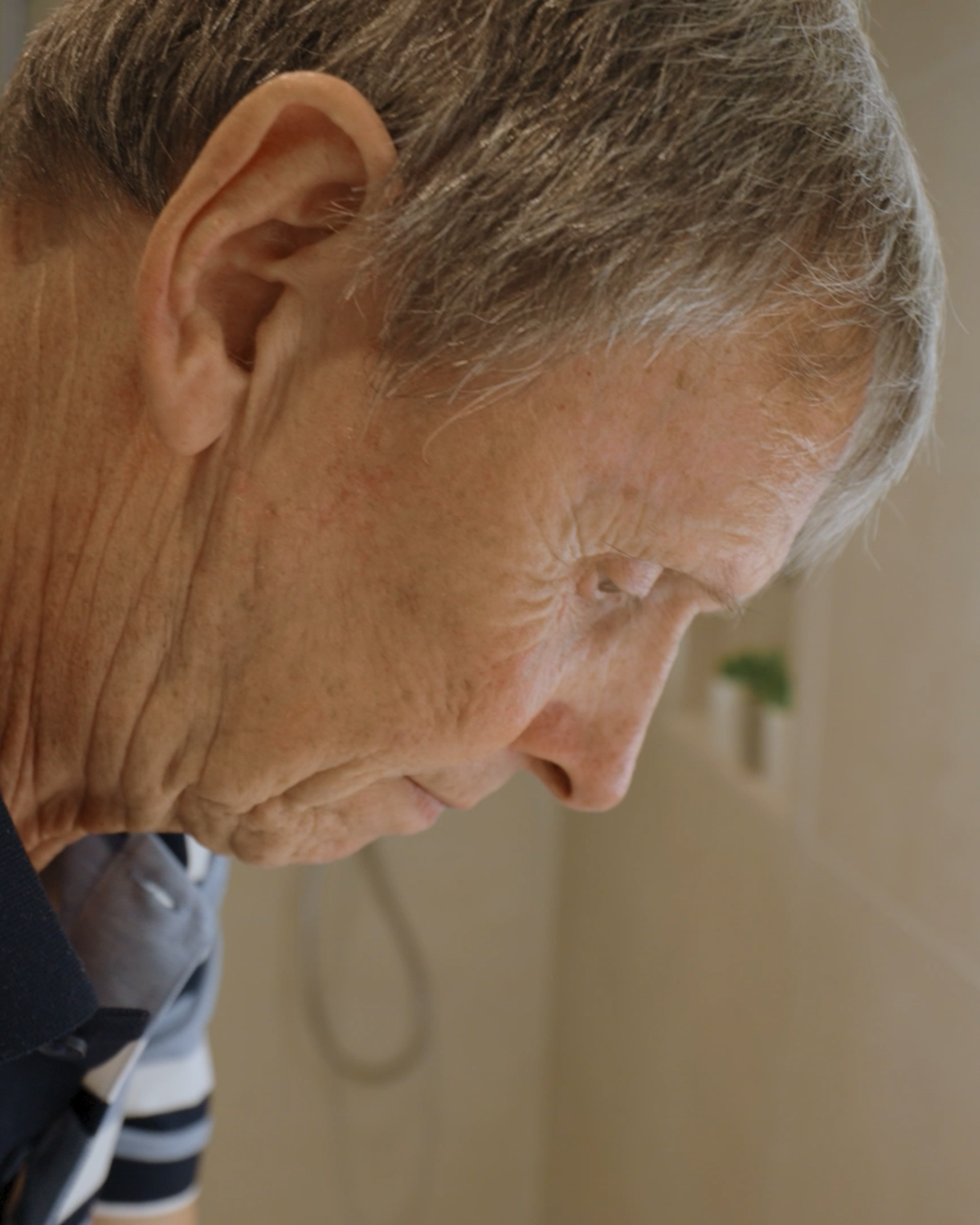 Elderly man with gray hair and striped shirt looking down, standing near a softly lit bathroom mirror.