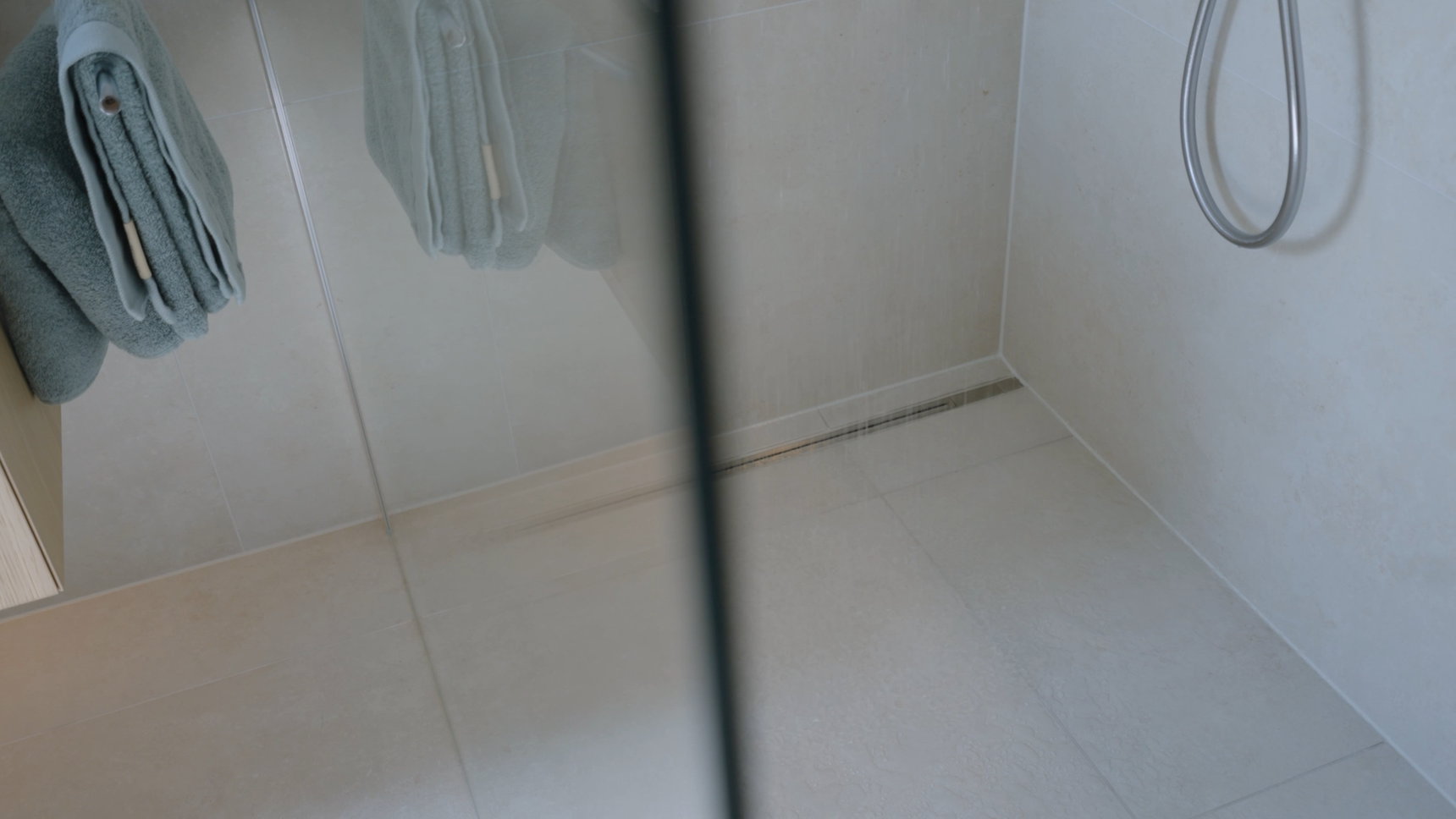 Minimalist shower with glass door, beige tiles, wall-mounted showerhead, and folded gray towels on a rack.