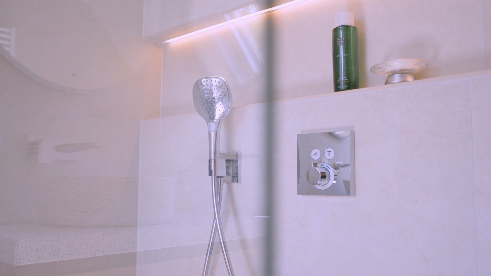 Modern shower with a silver showerhead, green bottle, and control panel on beige tiled walls, illuminated by soft lighting.
