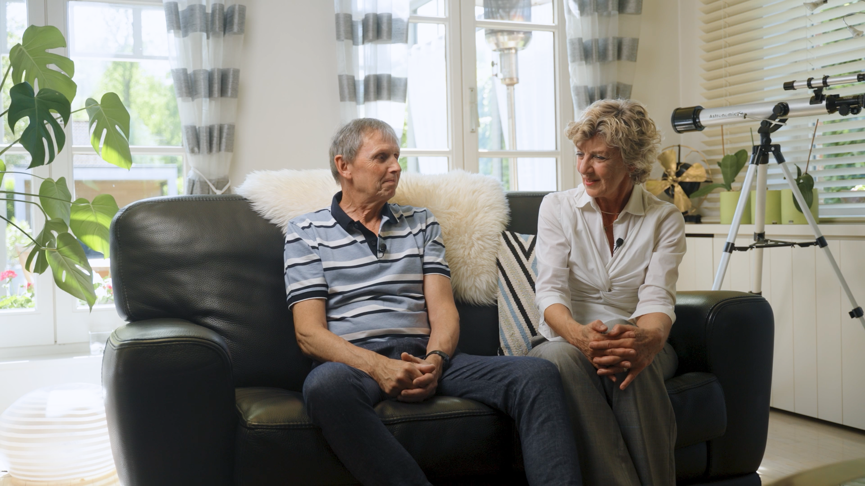 An older man and woman sit on a dark sofa in a well-lit living room, engaged in conversation. A telescope is visible near the window.