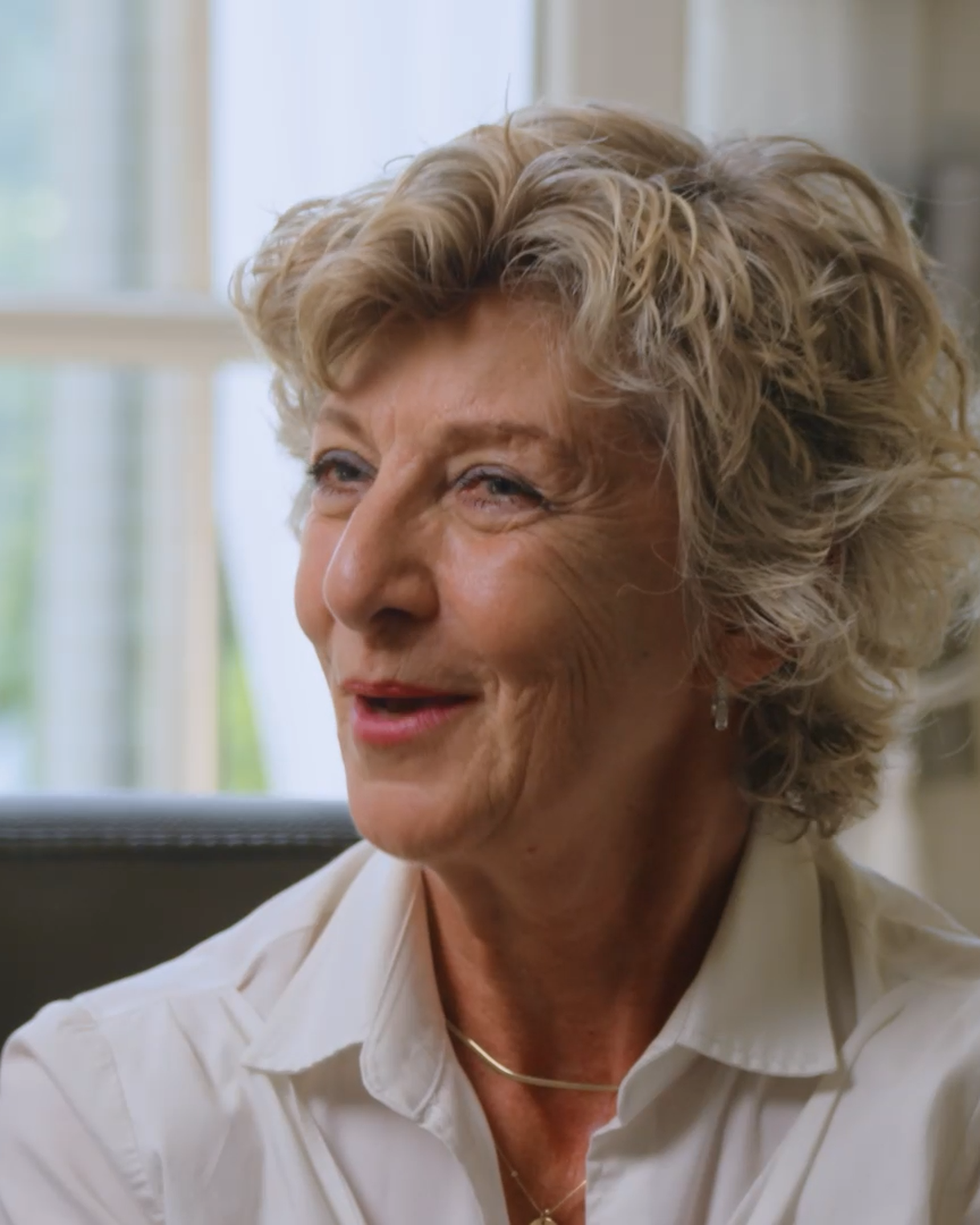 Smiling older woman with curly hair in a white shirt, sitting indoors near a window with soft natural light.