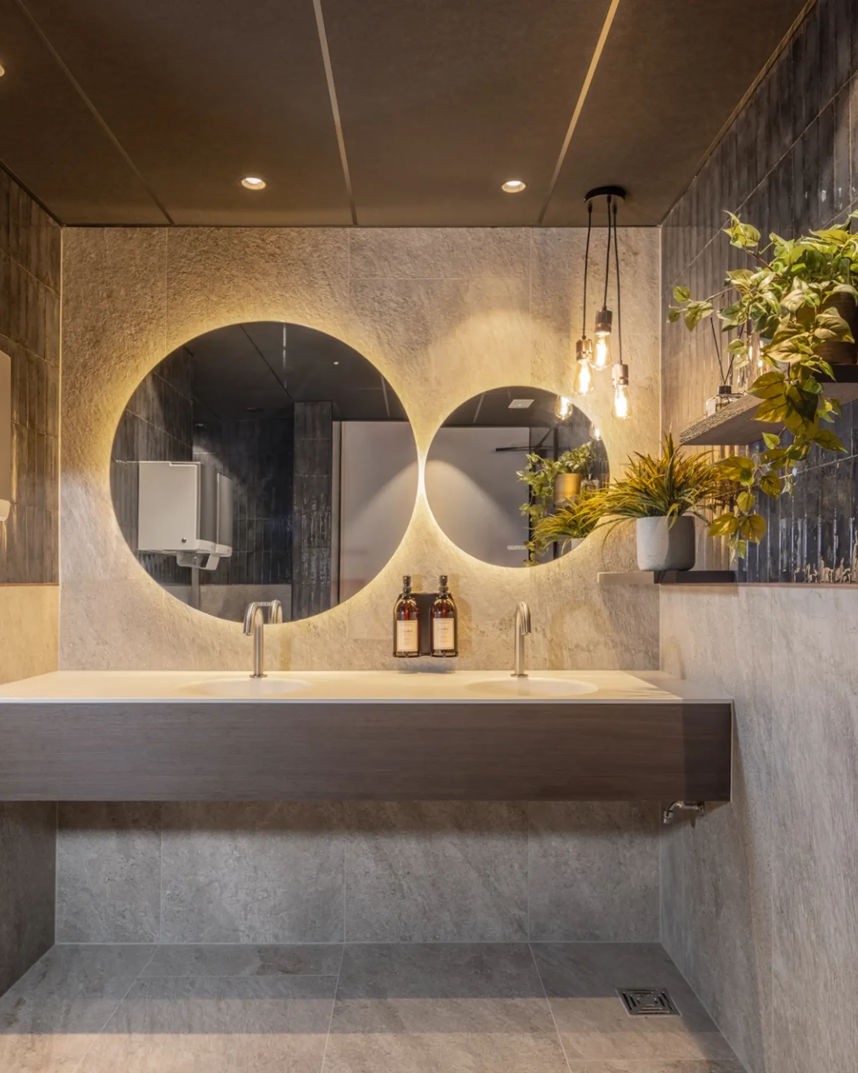 Modern bathroom with round mirrors, sleek sink, and hanging plants. Dim lighting and textured walls create a warm, inviting ambiance.