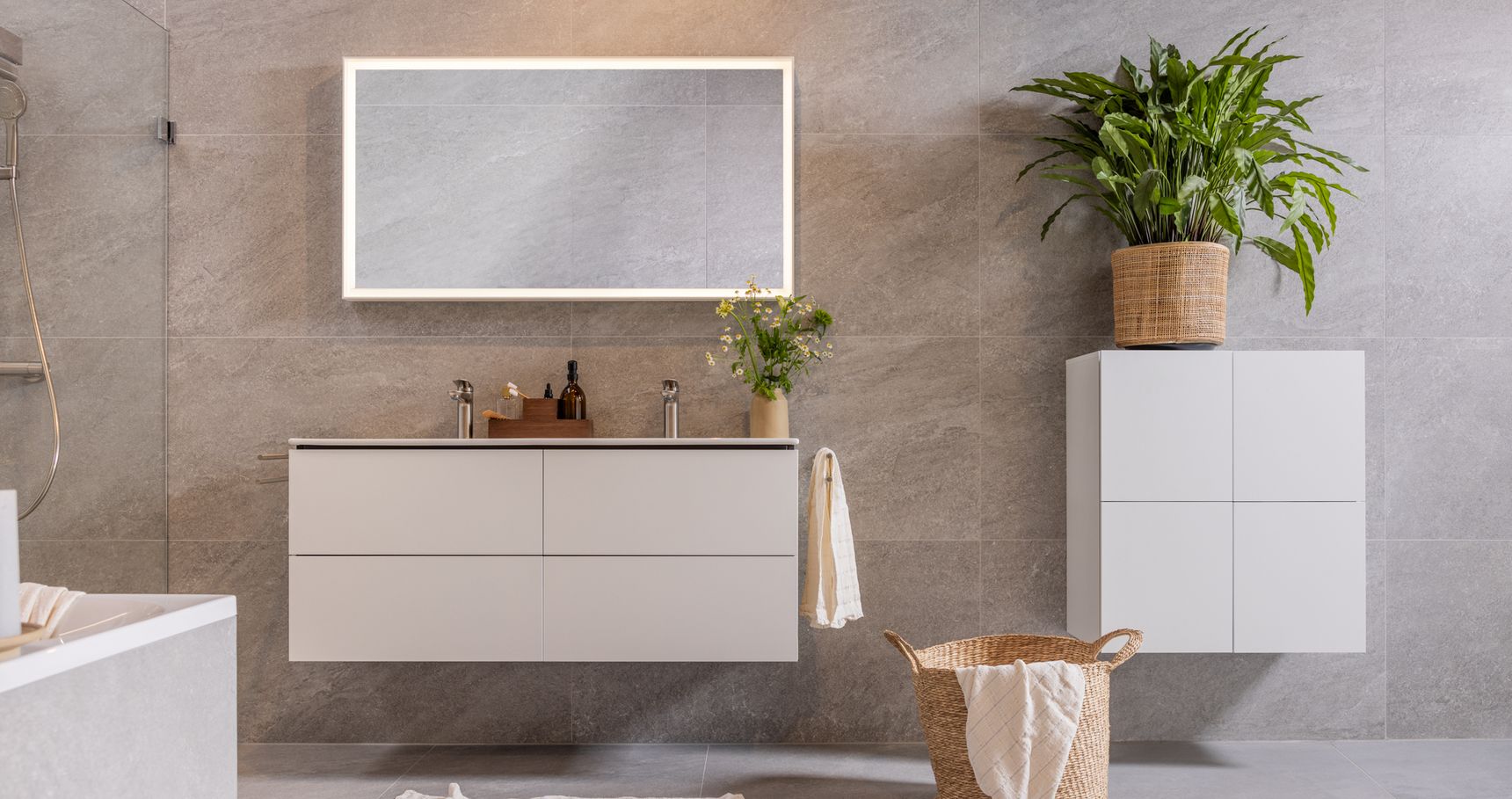 Moderne badkamer met zwevend badkamermeubel, verlichte spiegel, kamerplanten en rieten wasmand op een grijze tegelvloer.