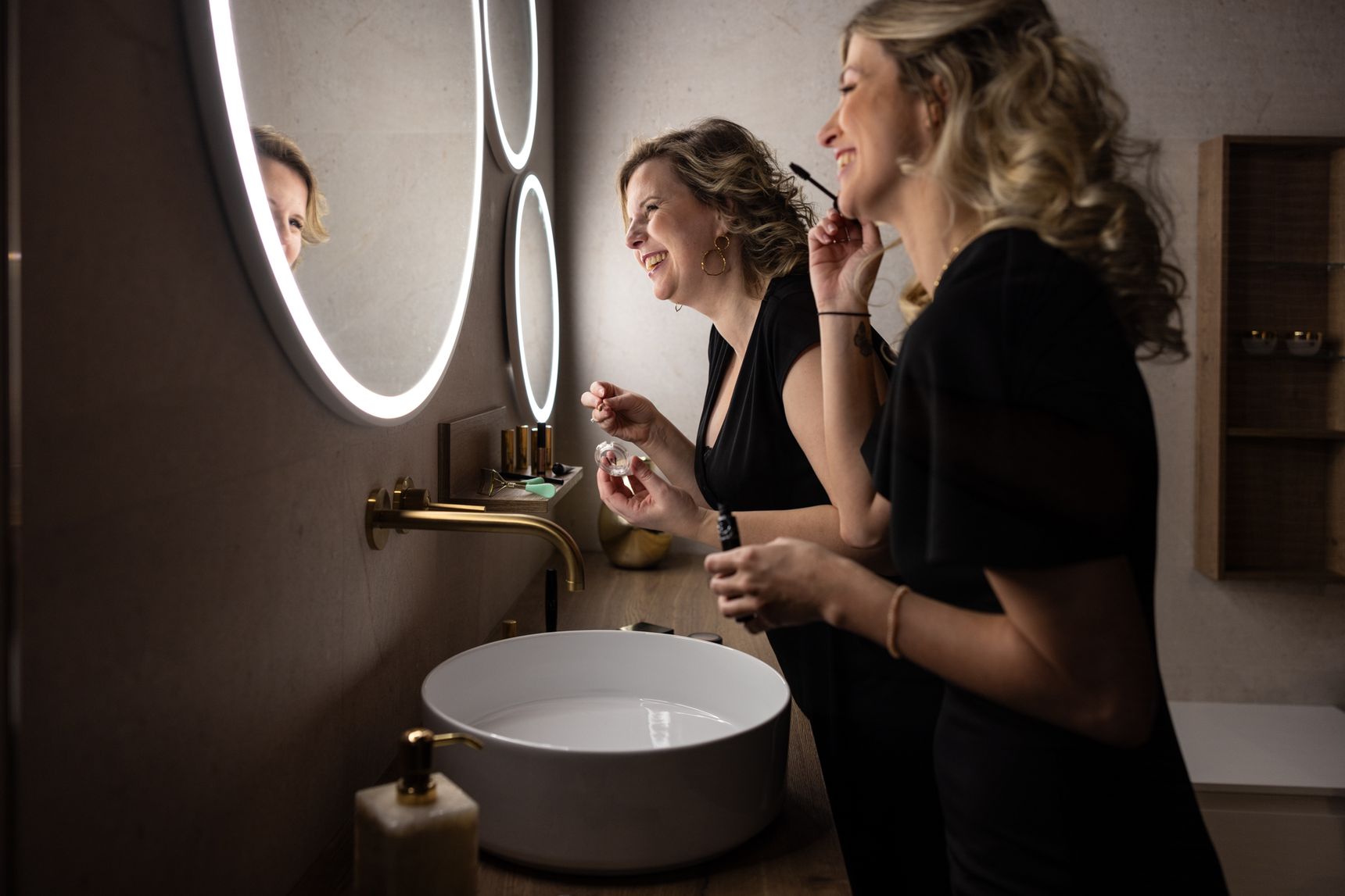 Twee vrouwen glimlachen en brengen make-up aan voor een ronde, verlichte spiegel in een moderne badkamer.