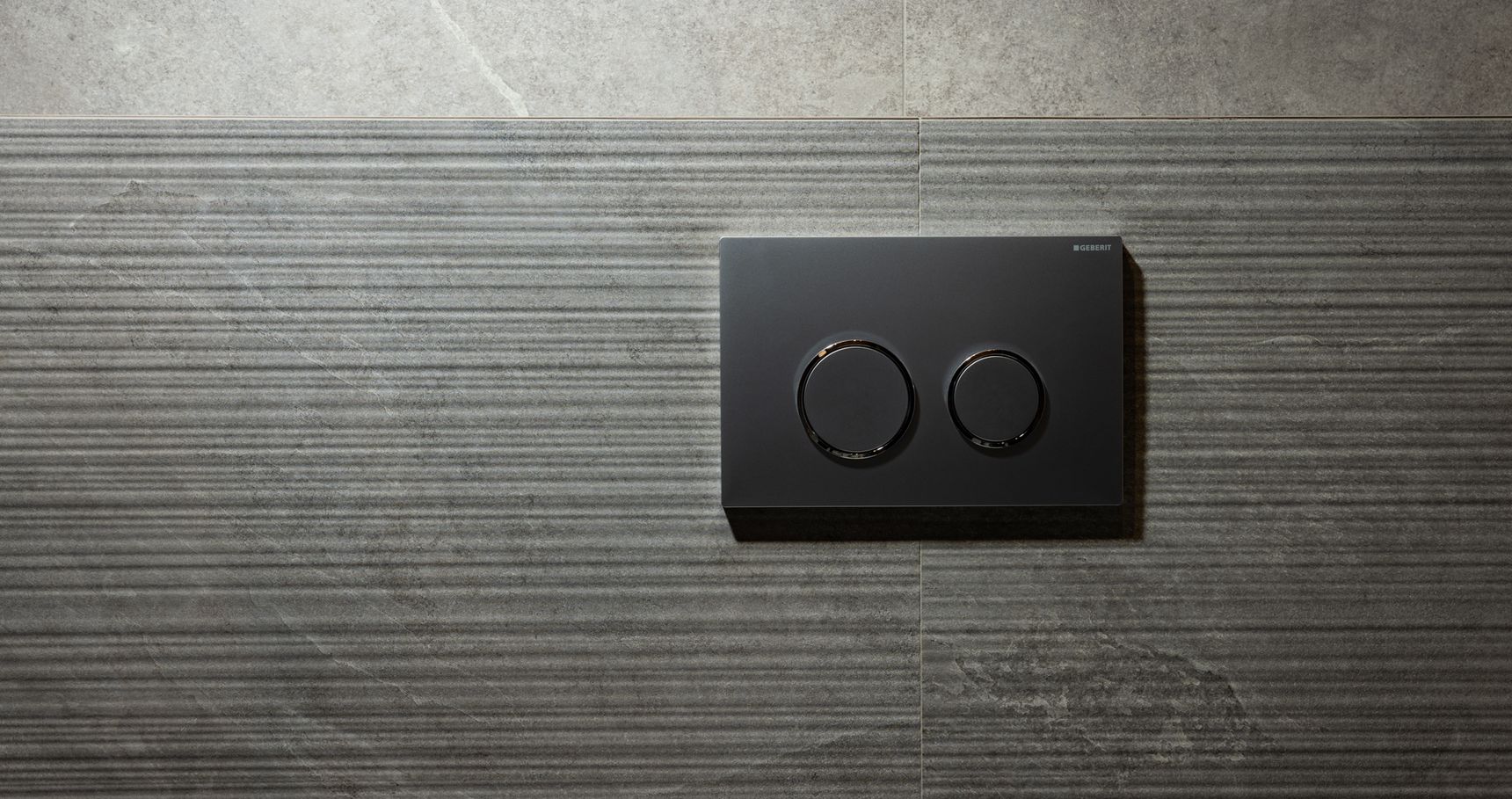 Modern bedieningspaneel met dual-flush systeem en twee ronde knoppen op een getextureerde grijs betegelde wand, voor waterbesparend doorspoelen.