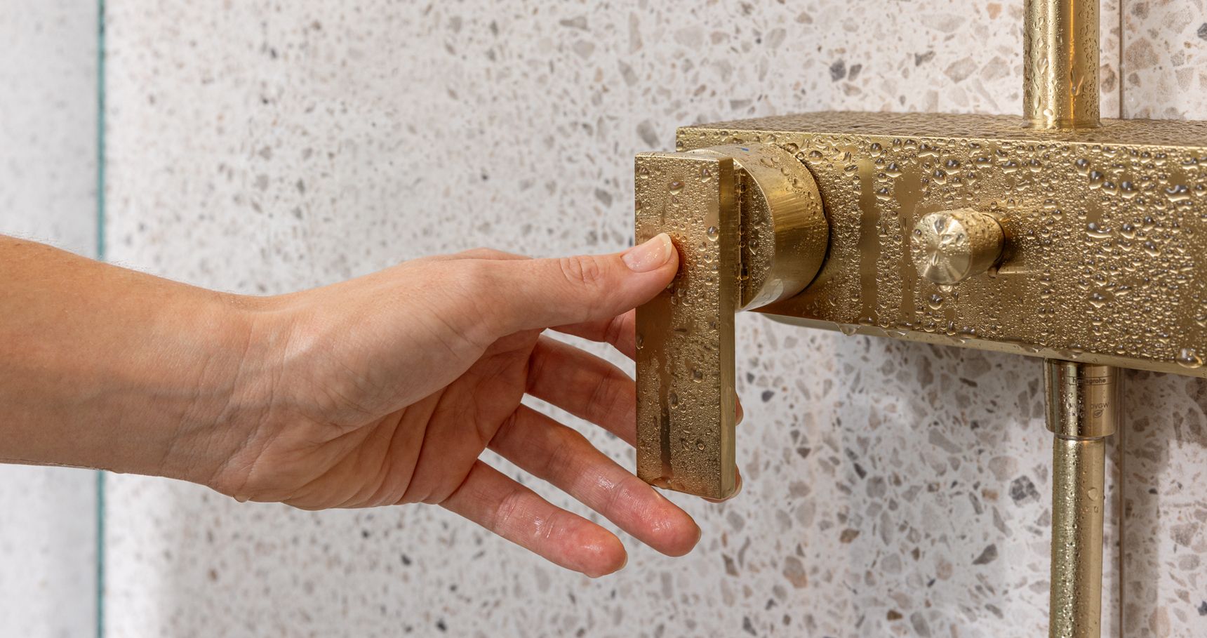 Hand stelt natte goudkleurige douchekraan af tegen een gespikkelde tegelwand.