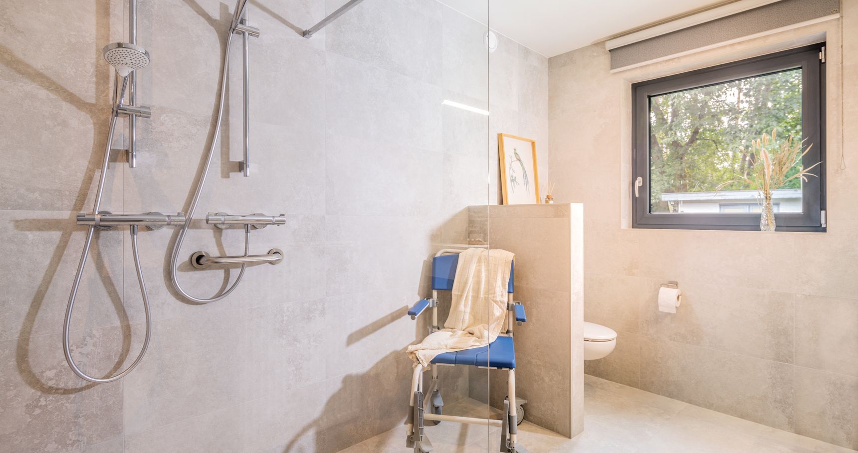 "Toegankelijke badkamer met inloopdouche, steunbeugels, douchestoel en raam, met lichtgekleurde tegels en handdoek over de stoel.