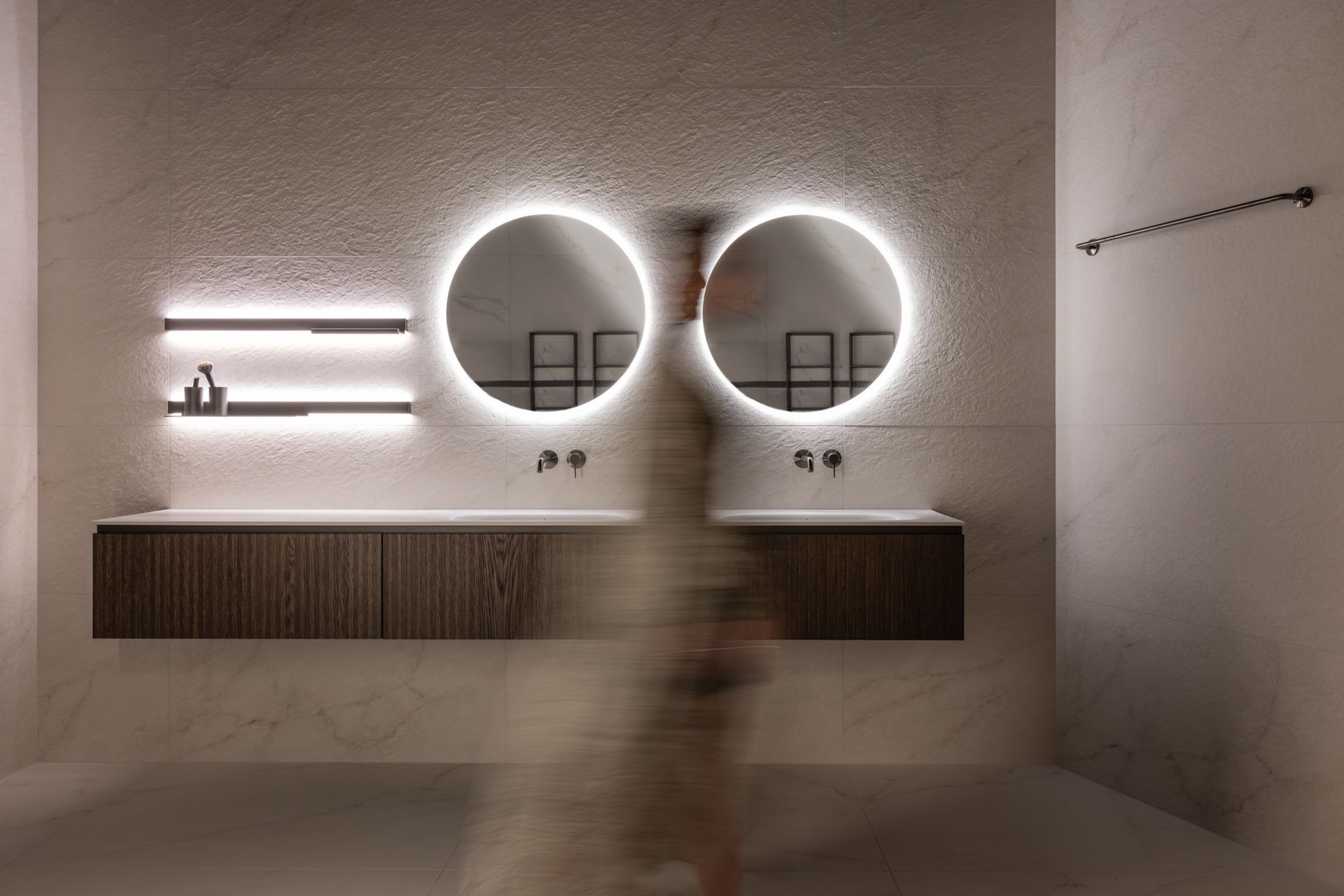 Moderne badkamer met twee verlichte ronde spiegels, houten dubbel badkamermeubel en een vervaagde persoon die voorbij wandelt, in een minimalistisch en elegant interieur.