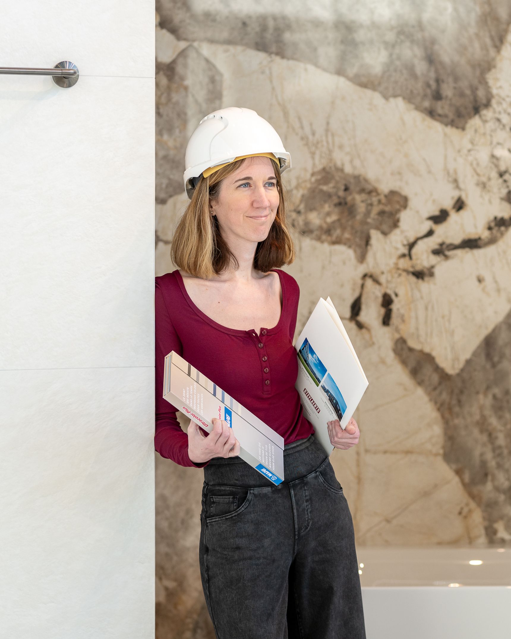 Woman in a hard hat holding tile samples, standing by a marble-patterned wall and a bathtub, wearing a burgundy top and jeans.