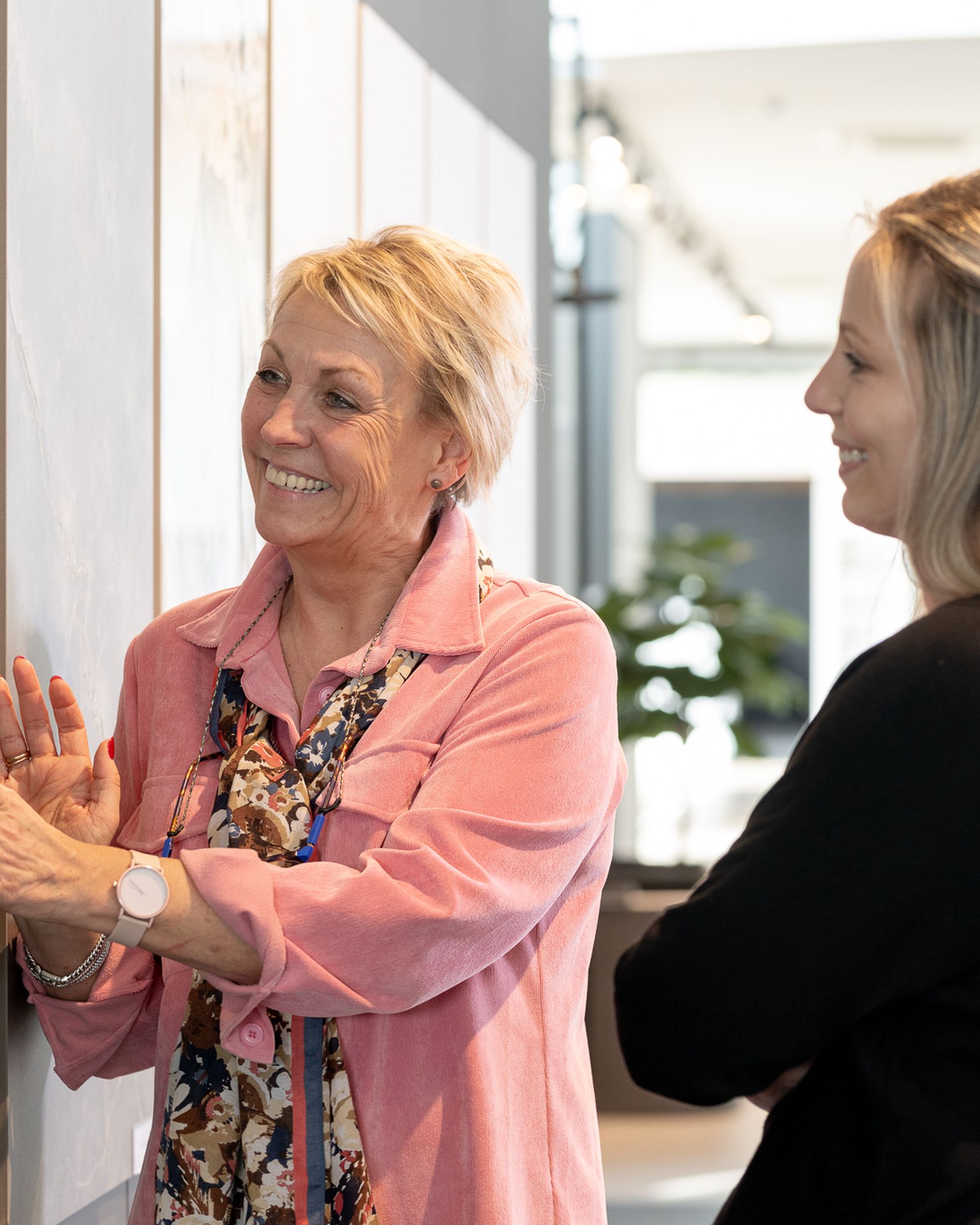 Two women smiling and looking at a wall display in a bright, modern gallery. One woman is touching the surface, while the other observes.