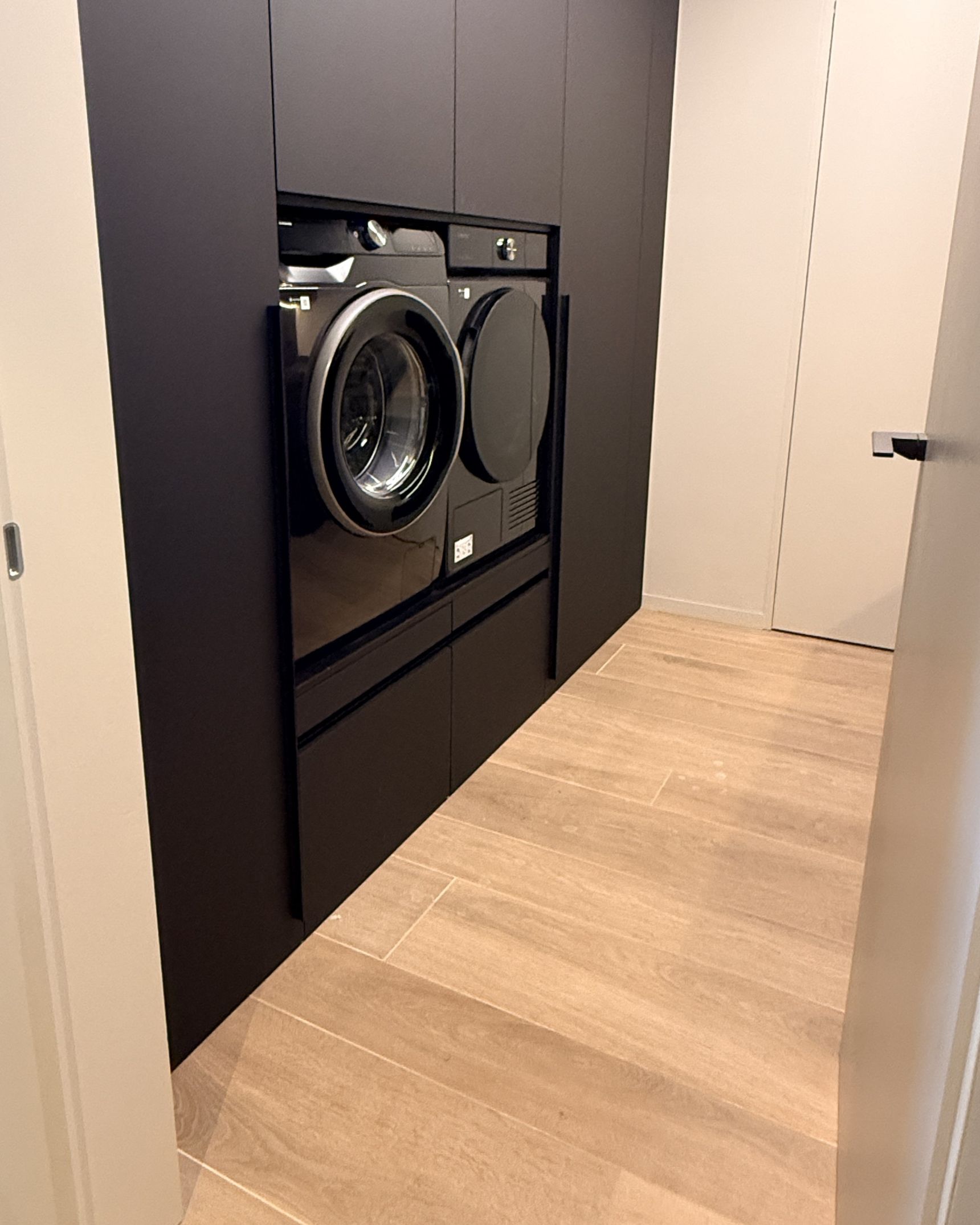 Ingebouwde wasmachine en droogkast in een strakke, donkere kast met houtlook vloertegels in een moderne wasruimte.