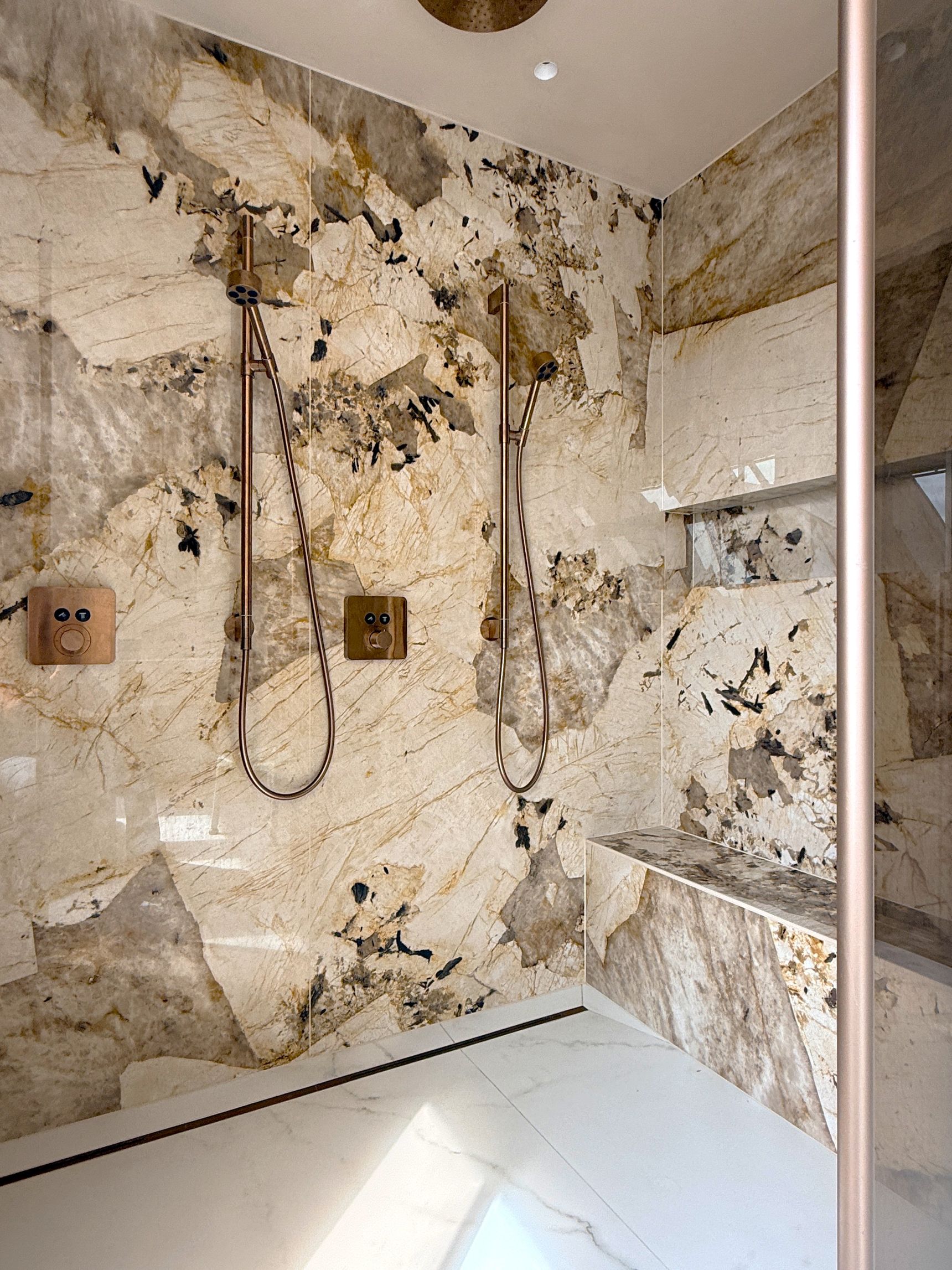 Inloopdouche met wanden in beige-bruine natuursteenlook, dubbele koperkleurige handdouches en een marmeren zitbank.