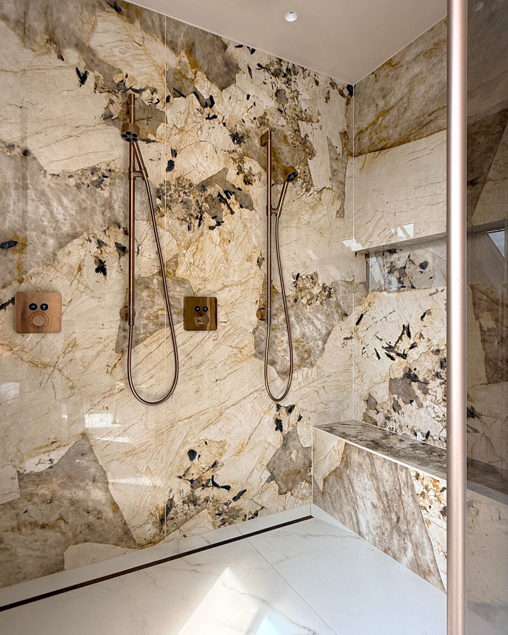 Inloopdouche met wanden in beige-bruine natuursteenlook, dubbele koperkleurige handdouches en een marmeren zitbank.