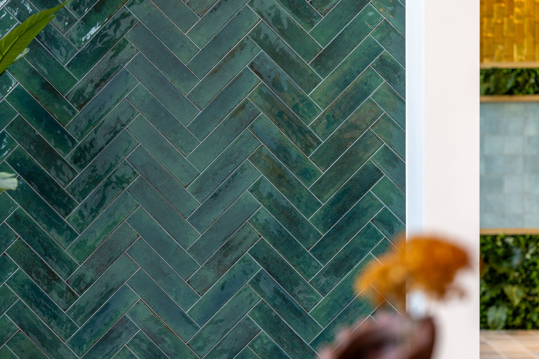 Close-up van een wand bekleed met glanzende groenblauwe keramische tegels in visgraatverband. De tegels tonen subtiele kleurvariaties en een ambachtelijke uitstraling. Op de voorgrond zijn onscherpe planten zichtbaar, terwijl op de achtergrond lichte architecturale elementen en groen zorgen voor een frisse, natuurlijke en eigentijdse sfeer.