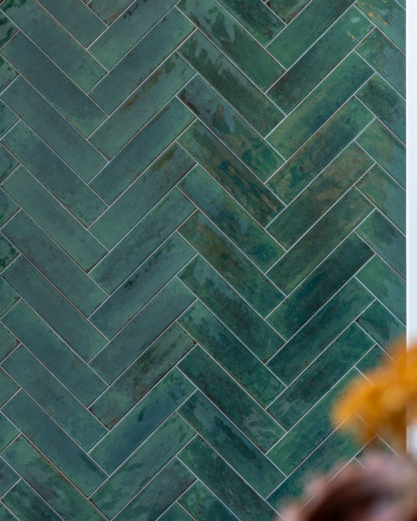 Close-up van een wand bekleed met glanzende groenblauwe keramische tegels in visgraatverband. De tegels tonen subtiele kleurvariaties en een ambachtelijke uitstraling. Op de voorgrond zijn onscherpe planten zichtbaar, terwijl op de achtergrond lichte architecturale elementen en groen zorgen voor een frisse, natuurlijke en eigentijdse sfeer.