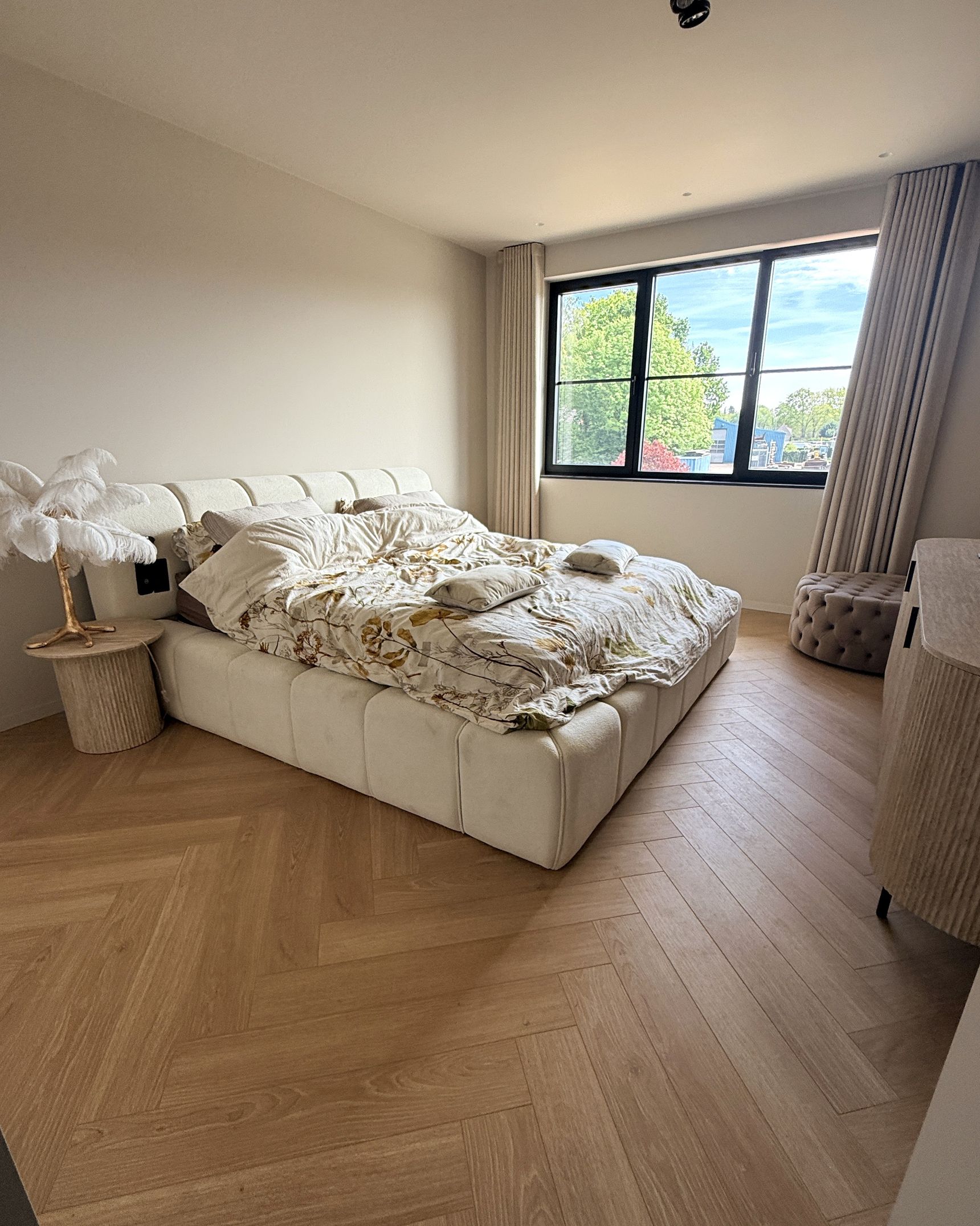 Slaapkamer met gestoffeerd crème bed, visgraatvloer in houtlook en groot raam met zicht op groen.