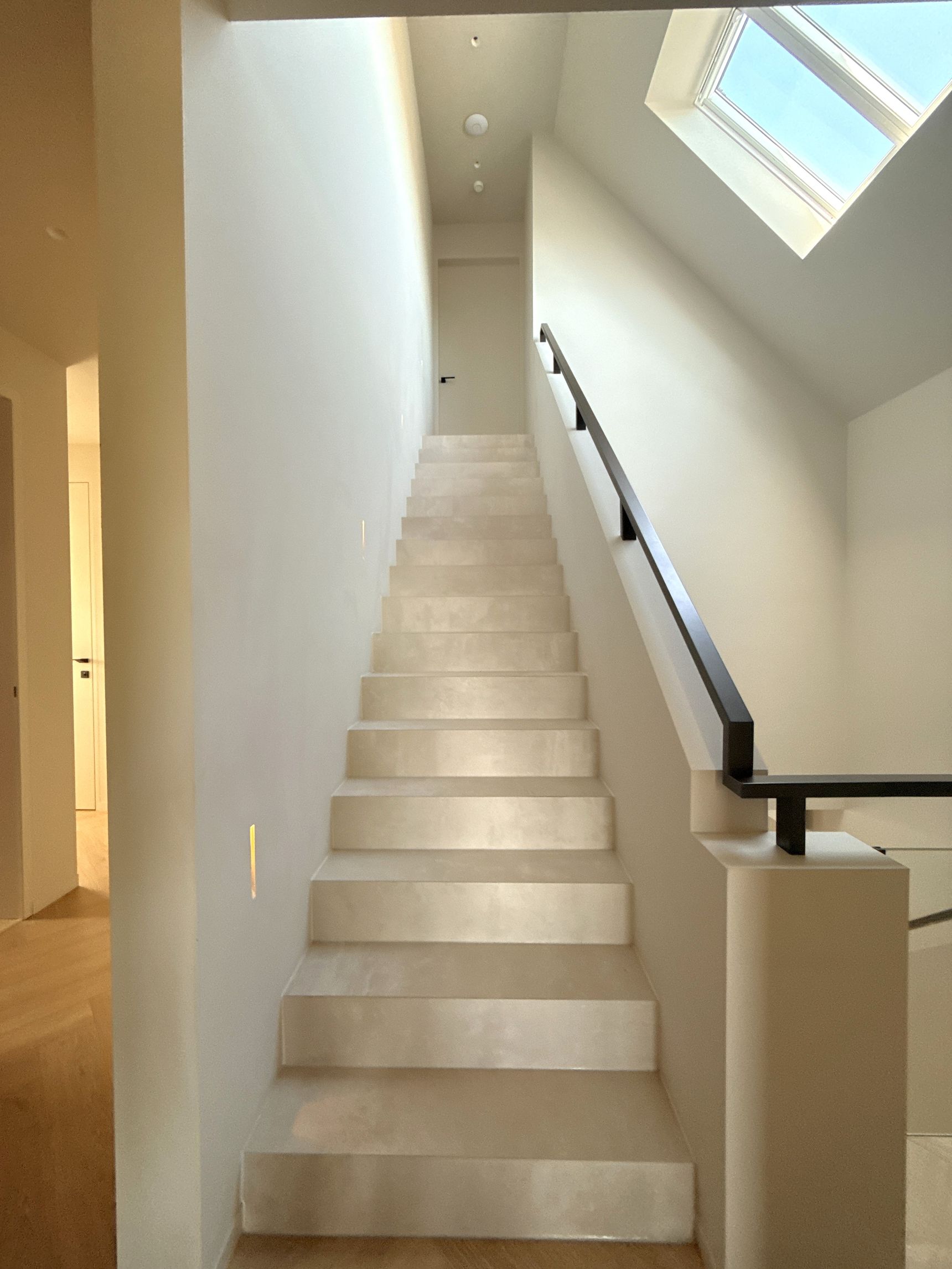 Trap met lichte betonlook treden, zwarte leuning en dakraam boven het trapgat.