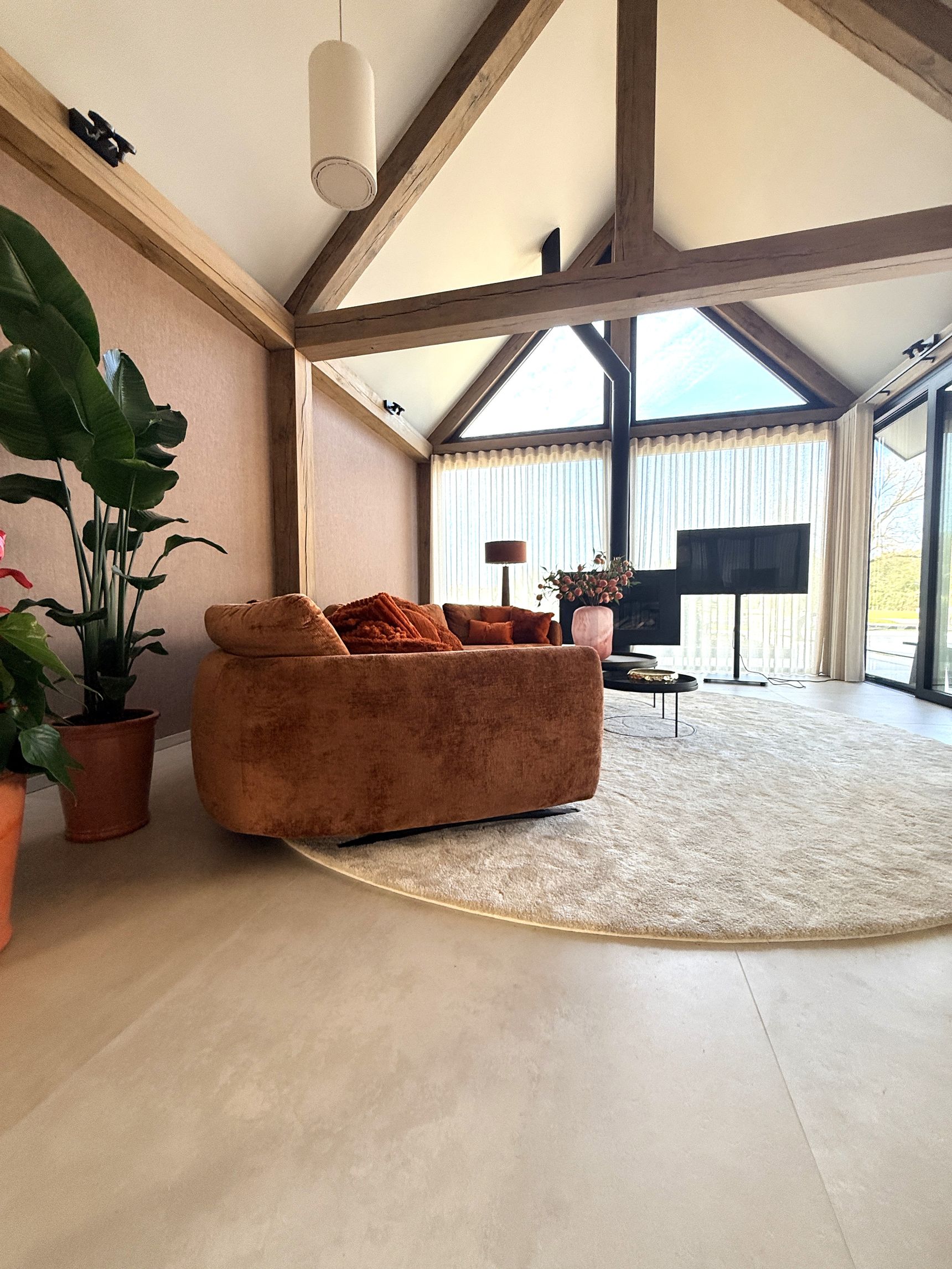 Zithoek met roestbruine bank onder houten dakspanten, ronde vloerkleed en driehoekig raam met tuinzicht.