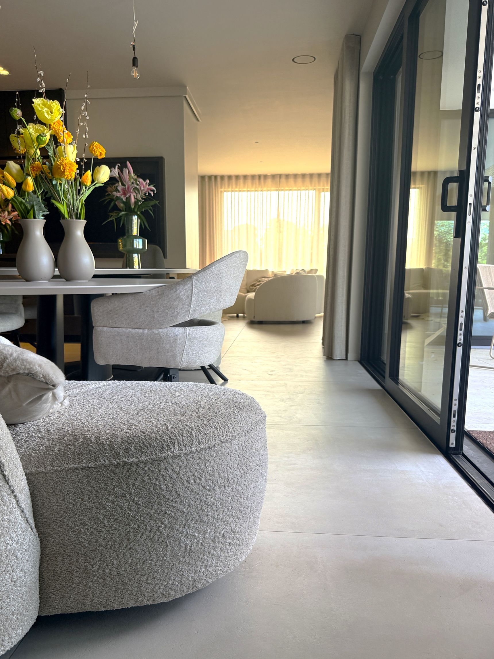 Woonkamer met bouclé zetel en poef, eettafel met bloemen en schuifdeuren naar het terras.