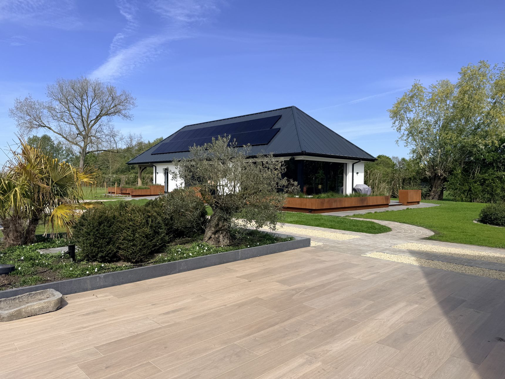 Moderne witte villa met zwart pannendak en zonnepanelen, omringd door een strak aangelegde tuin met houtlook terrastegels, olijfboom, siergrassen en een pad van kasseien en grind.