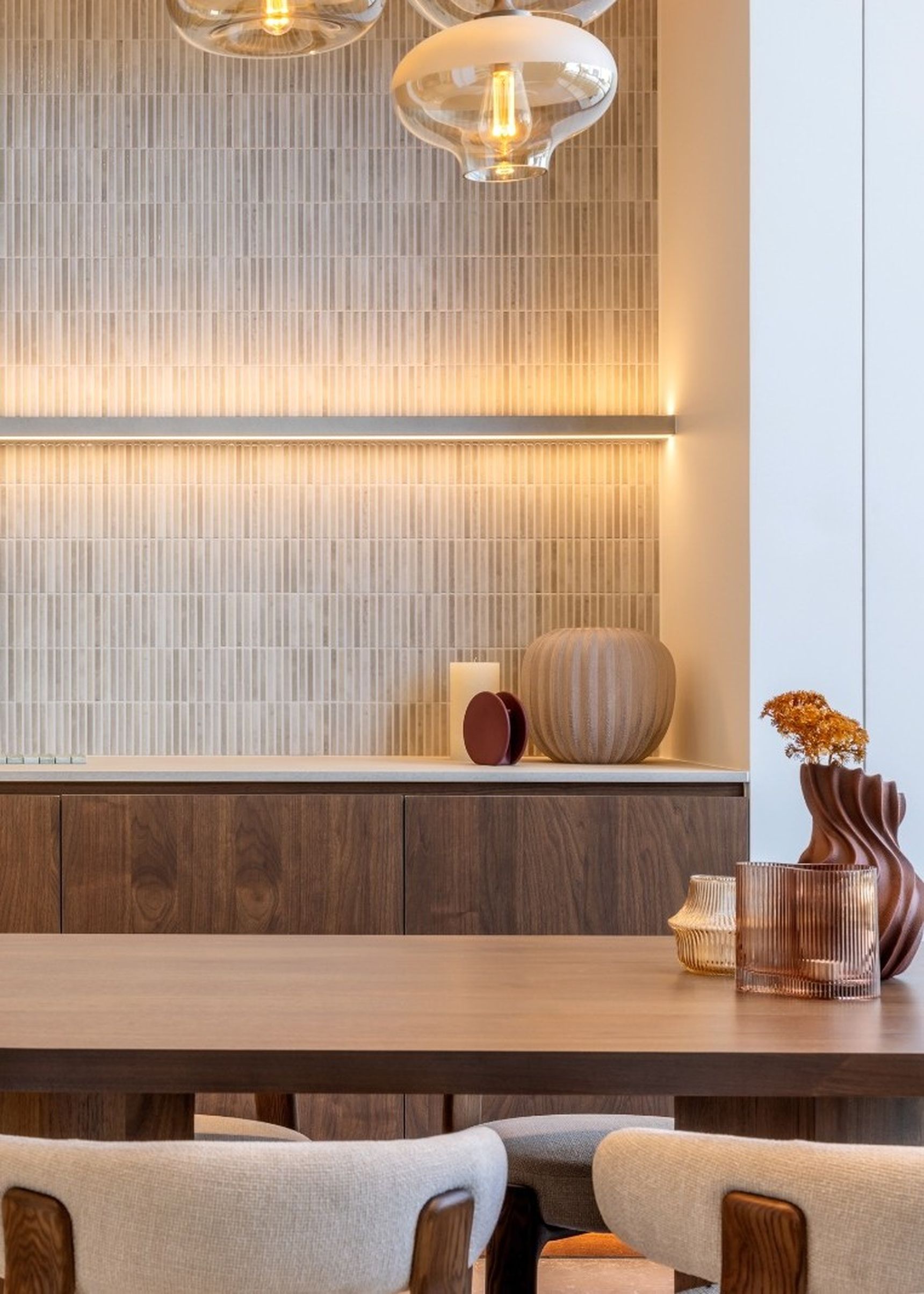 Warme, moderne eetruimte met maatwerk kasten in donker walnoothout, een wand met fijne verticale reliëftegels in zachte beige tinten en geïntegreerde ledverlichting. Op de voorgrond staan een ronde houten eettafel en gestoffeerde stoelen, terwijl glazen hanglampen en decoratieve accessoires bijdragen aan een gezellige, cocooning sfeer.