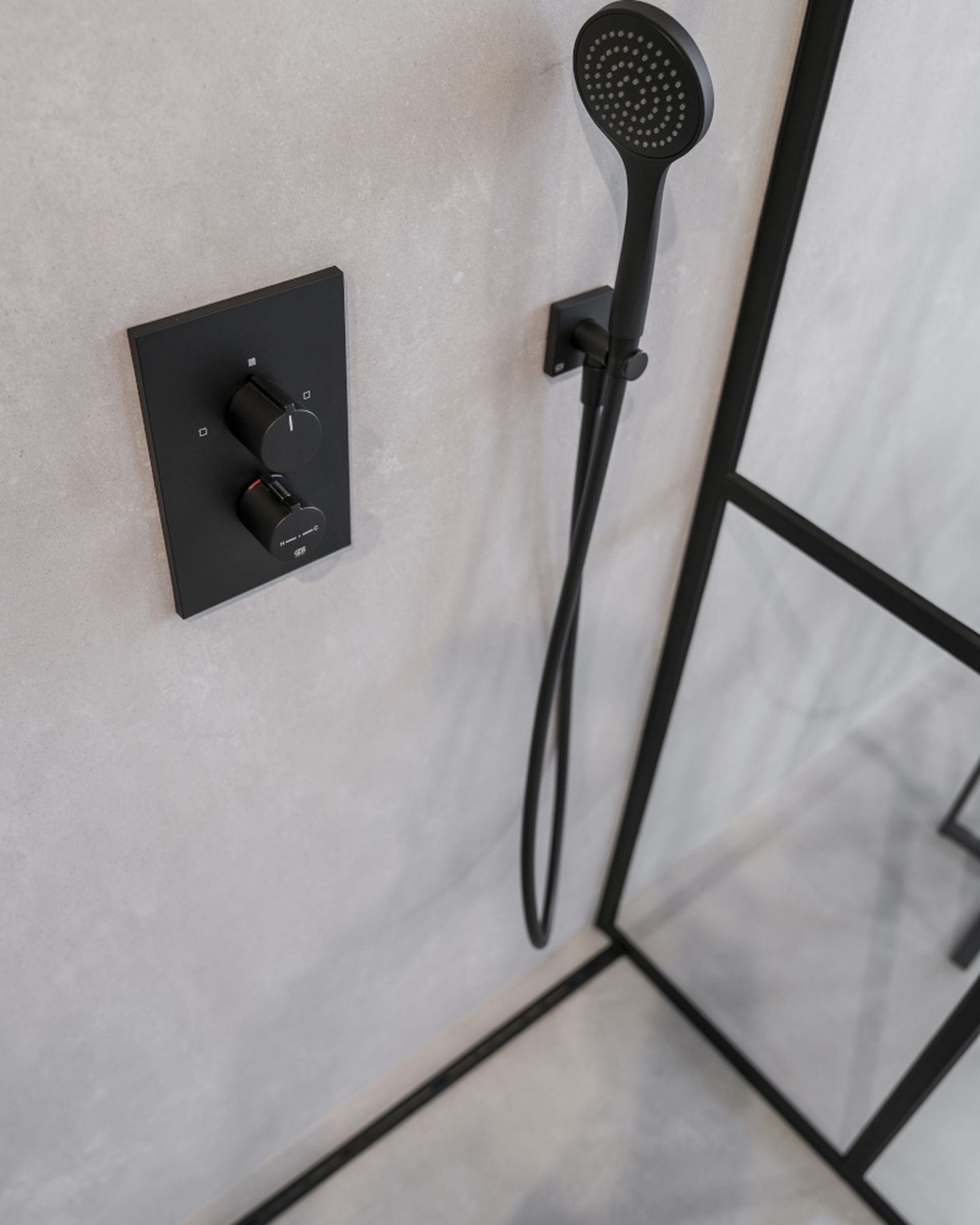 Modern shower with black handheld showerhead, black control panel, and glass partition on light gray tiled wall.