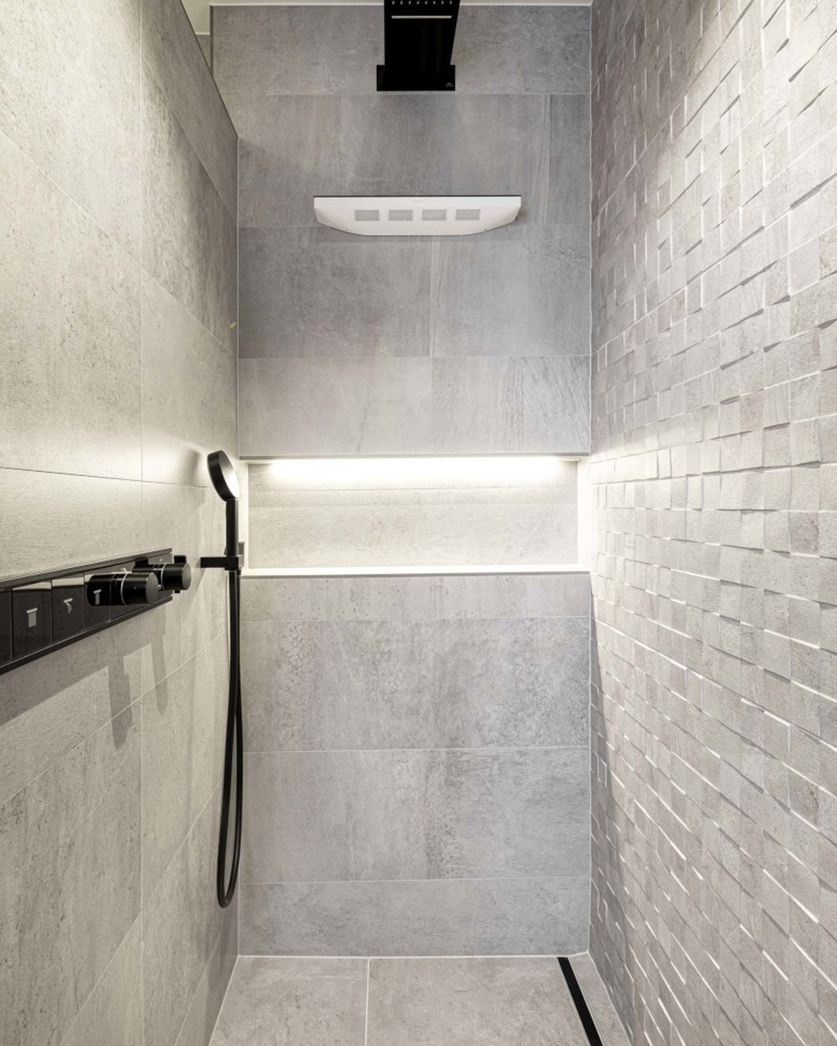 Modern shower with gray tiles, a black rainfall showerhead, and textured wall. LED strip lighting highlights the sleek design.