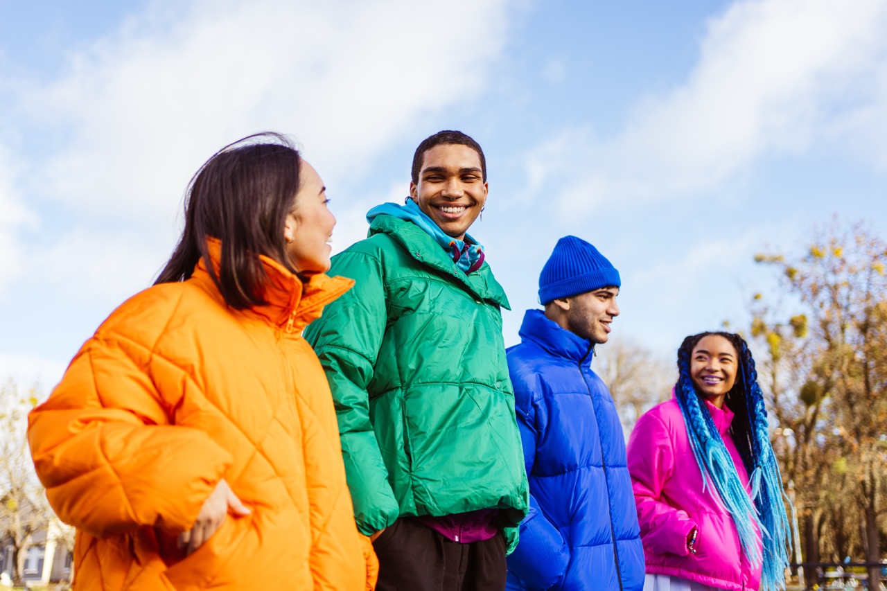 Vier mensen in kleurrijke puffer winterjassen lachen buiten op een heldere dag, met bomen en een blauwe lucht op de achtergrond.