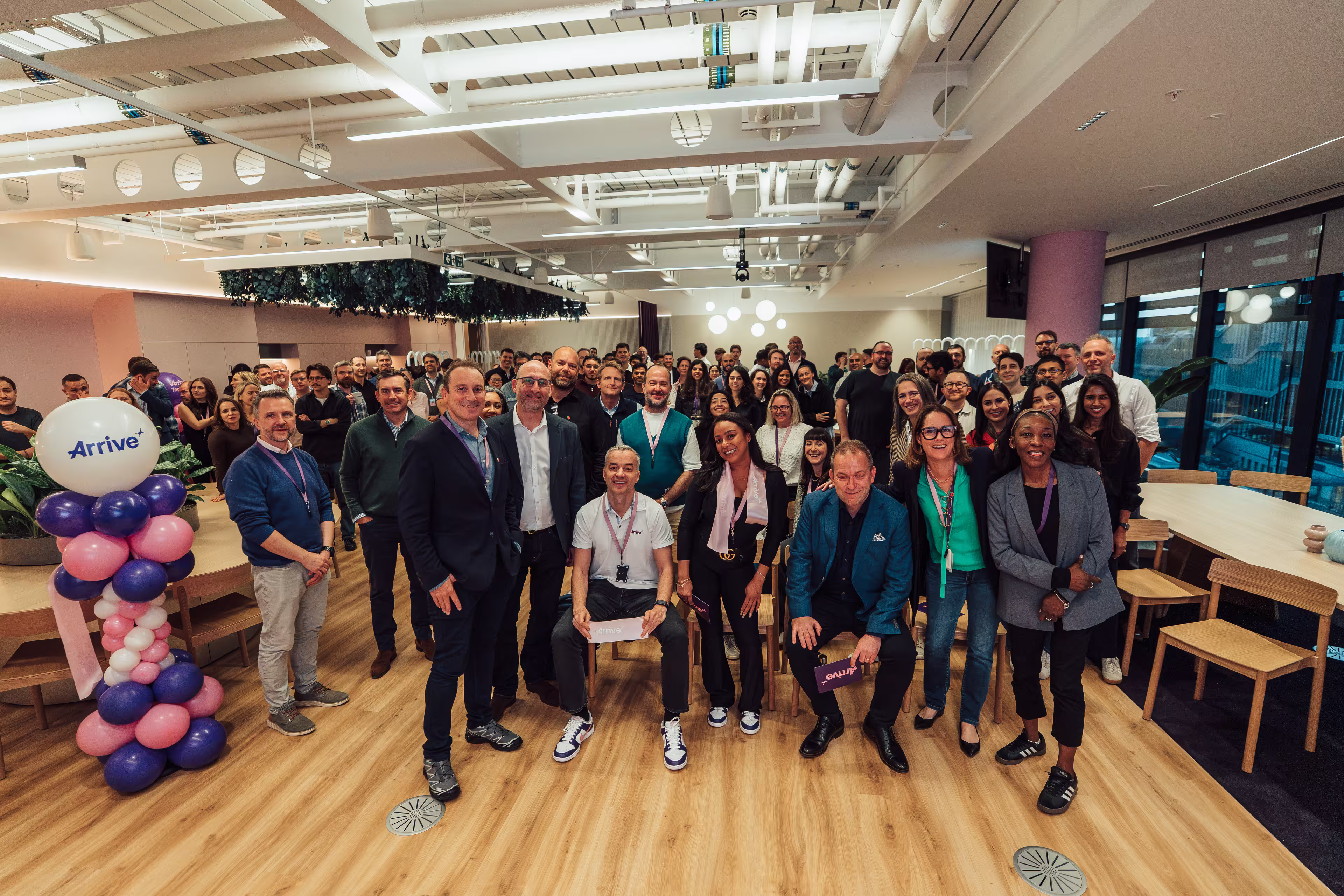 A group shot of people looking and smiling at the camera. To the left a balloon column with the Arrive logo at the top