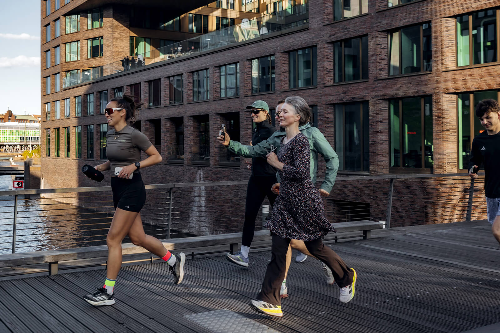 Vier Personen laufen lachend auf einem Holzsteg entlang eines Kanals in der Hamburger HafenCity, eine Teilnehmerin fotografiert mit dem Smartphone