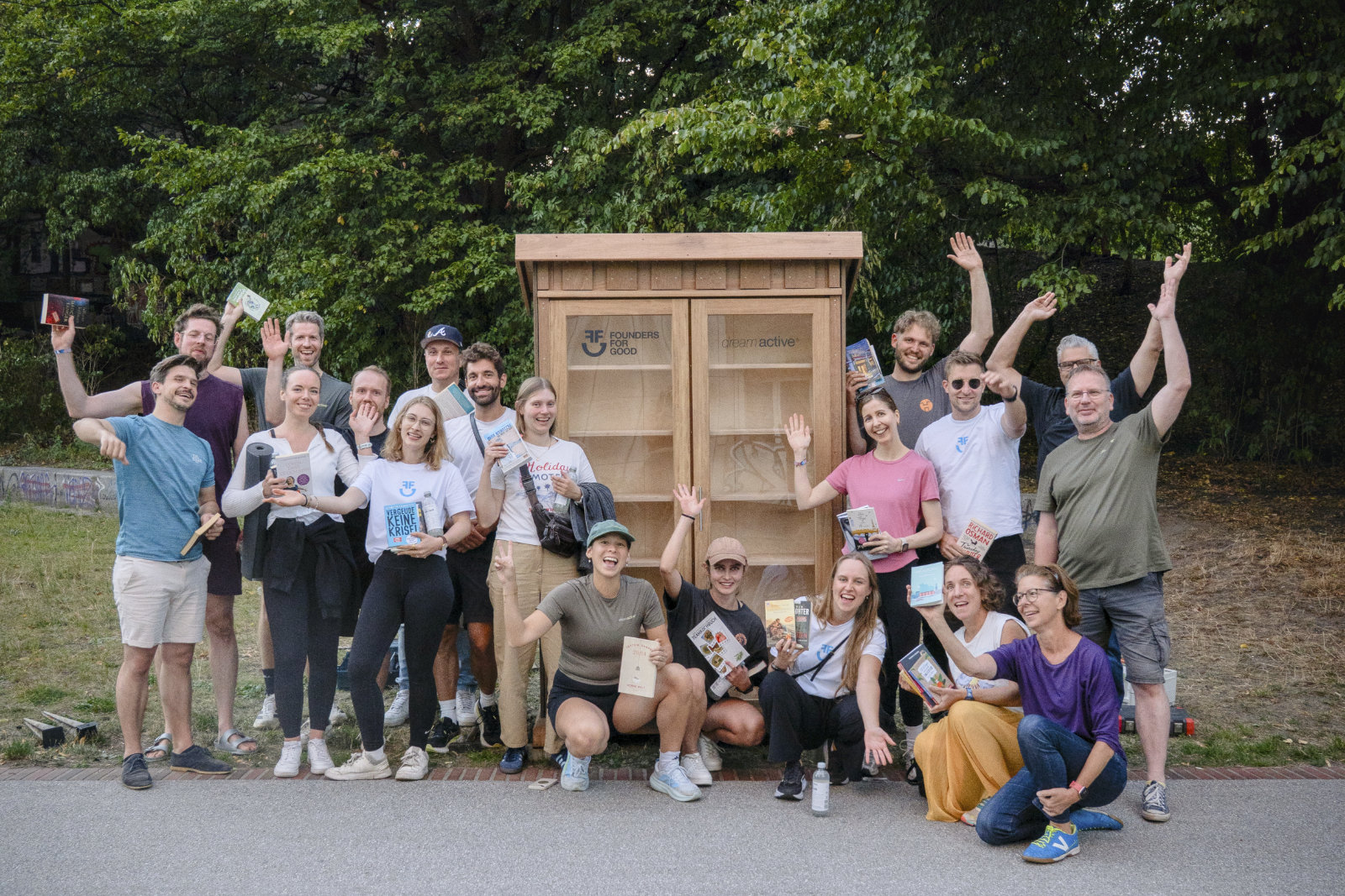 A group of people smiling and holding books, posing in front of a wooden book exchange cabinet in an outdoor area.