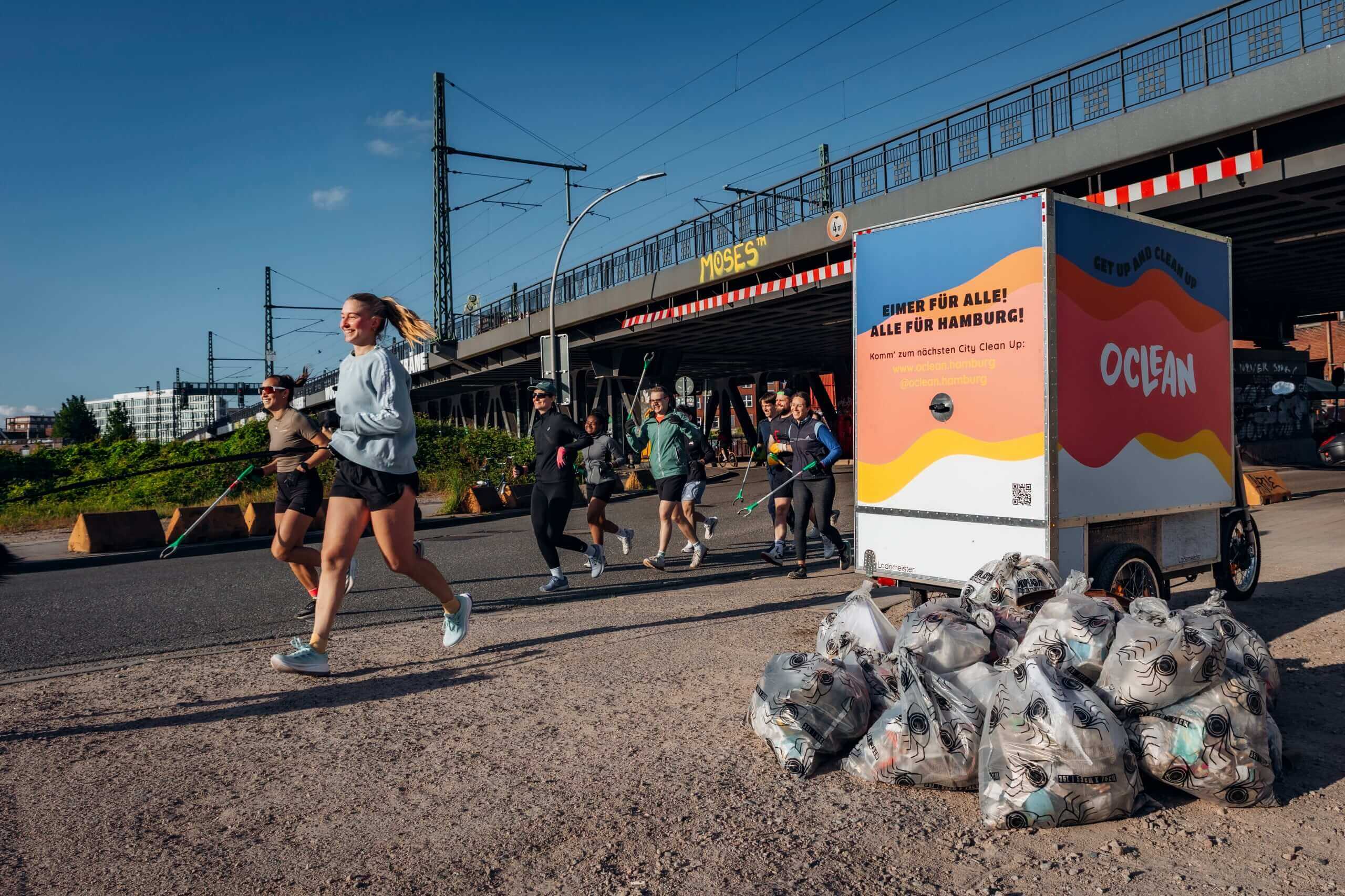 Laufgruppe mit Müllgreifern joggt lachend an einem Oclean-Anhänger vorbei, im Vordergrund zahlreiche gefüllte Müllsäcke des City Clean Ups in Hamburg
